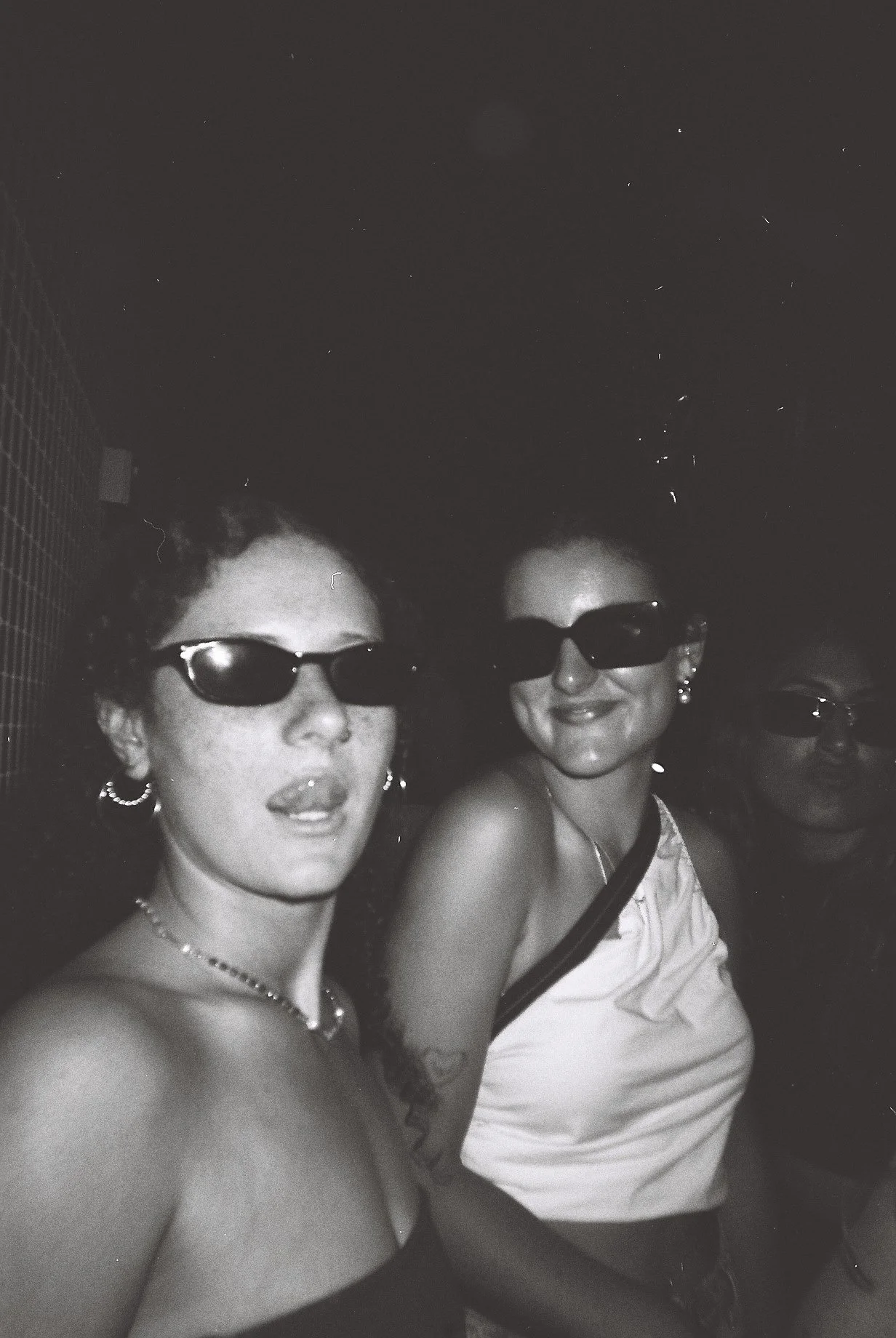 Two women wearing sunglasses smiling and posing for a photo in a dark setting, with a third person partially visible behind them.
