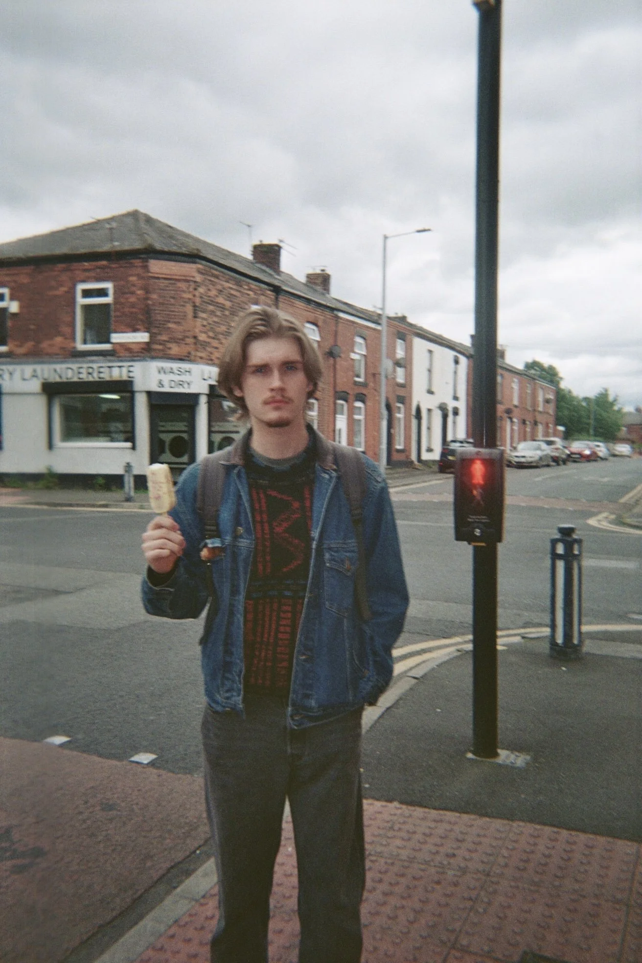 A young man with shoulder-length hair, standing on a city sidewalk, holding a popsicle, wearing a denim jacket over a black and red patterned shirt, with a backpack. The background shows residential buildings, parked cars, and a pedestrian crossing s