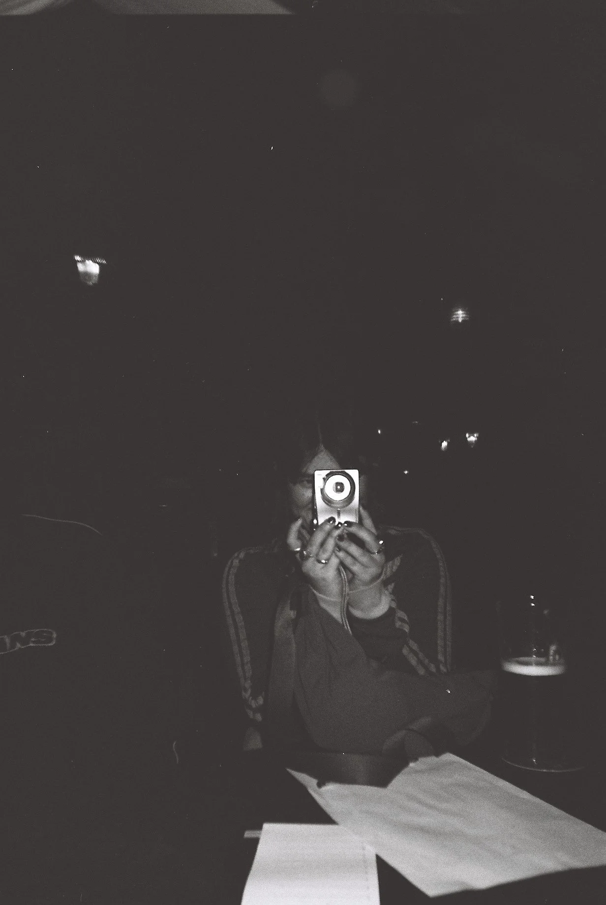 A person taking a selfie with a camera in a dimly lit room. They are sitting at a table with papers and a mug on it.