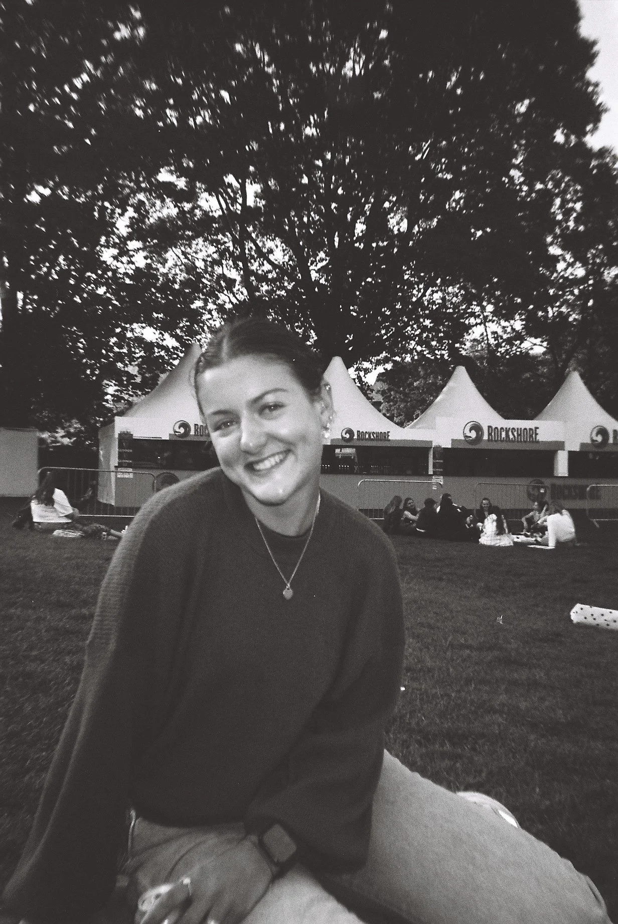 Black and white photo of a smiling woman sitting outdoors at night, with tents labeled "Rockshore" and a group of people in the background at an outdoor event.