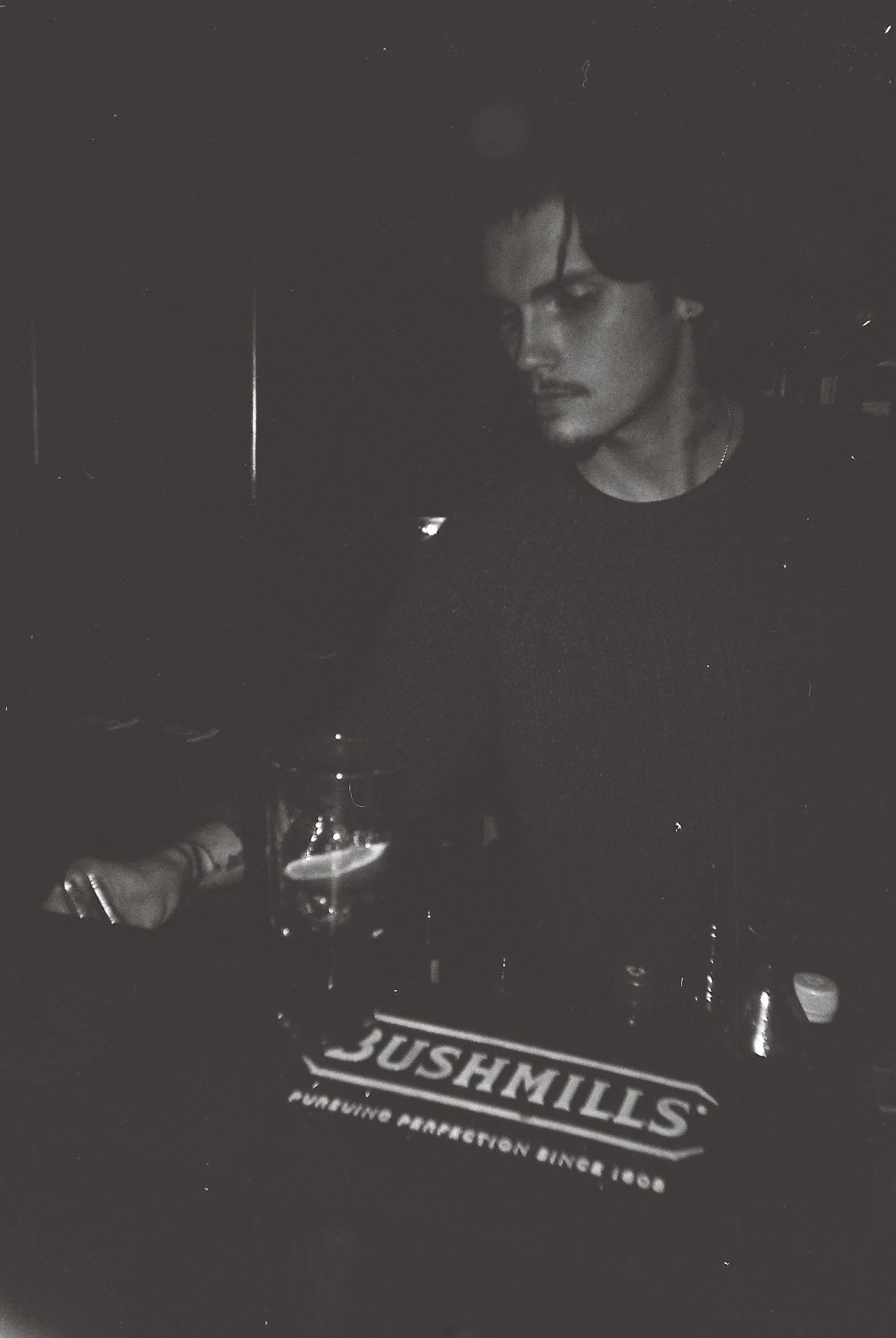 A person with dark hair and makeup sitting at a table in a dimly-lit environment, with a glass of drink in front of them and a badge reading "BUSHMILLS" on the table.