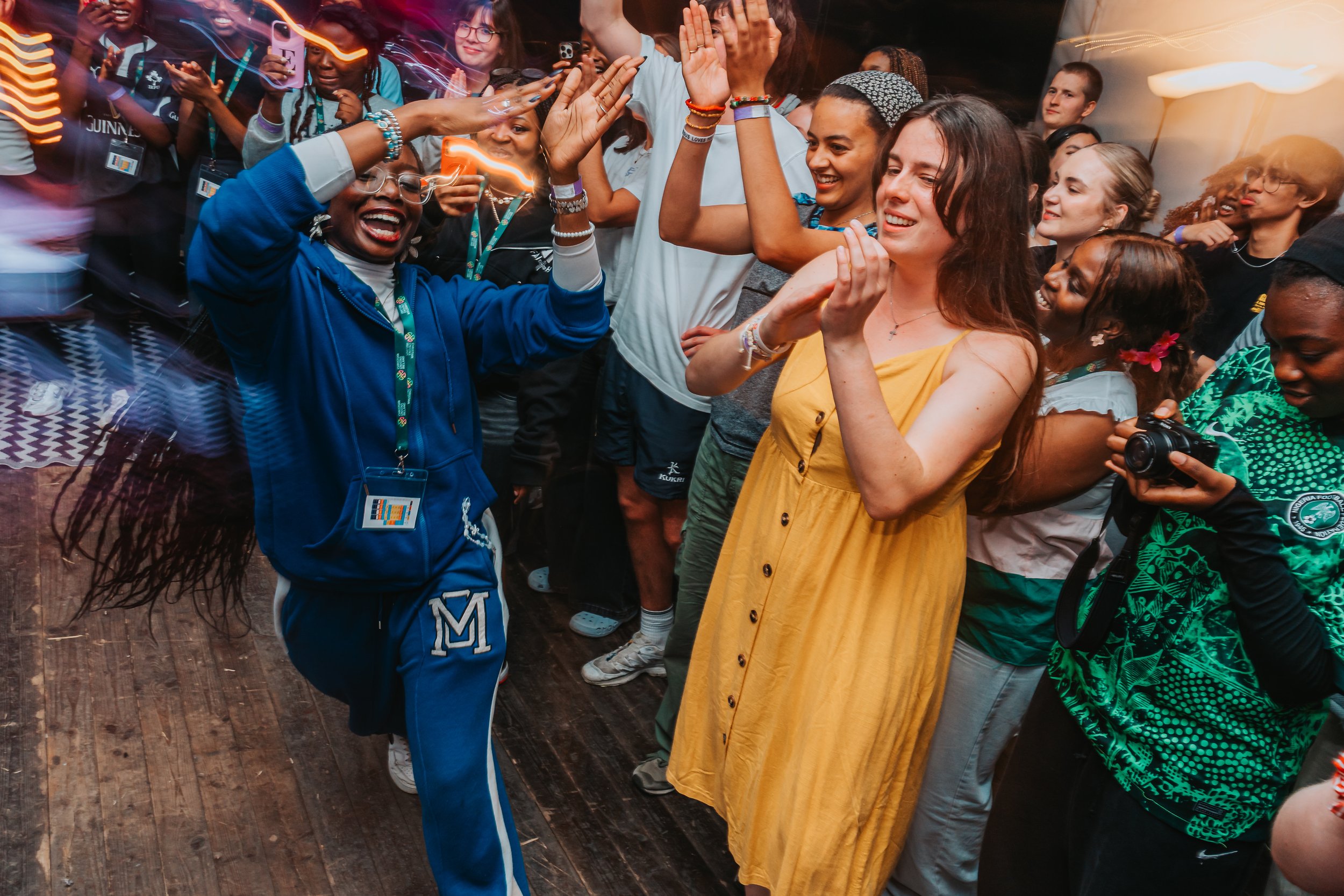 A diverse group of people dancing and celebrating at an indoor event, smiling, with some raising their hands and others taking photos.