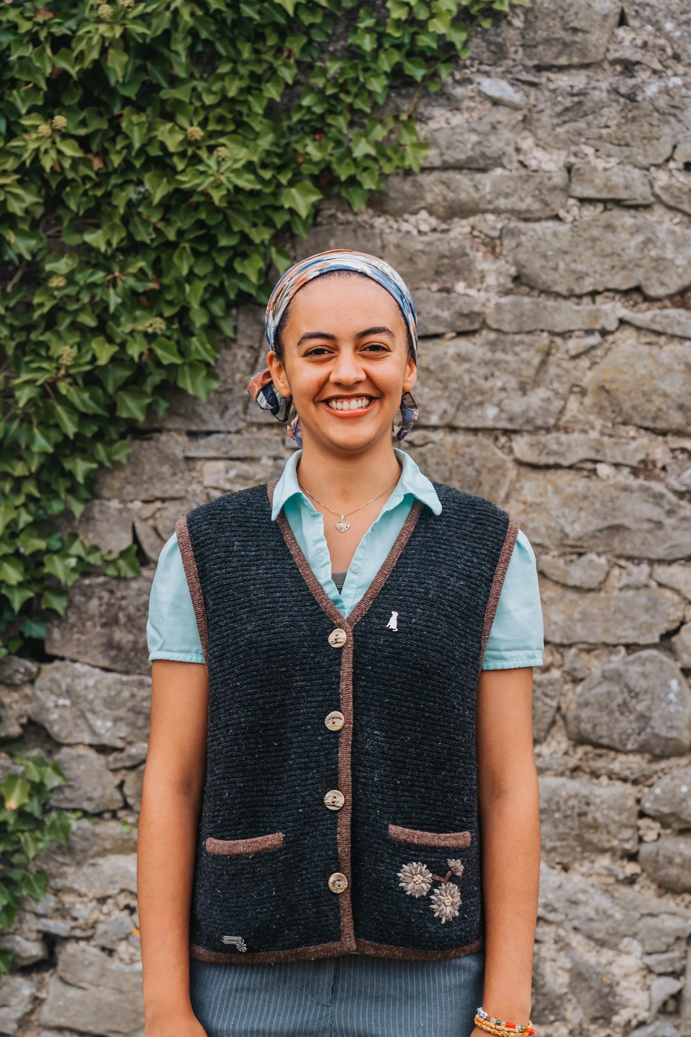 Portrait of a smiling young woman with a headscarf standing outdoors in front of a stone wall and green ivy.