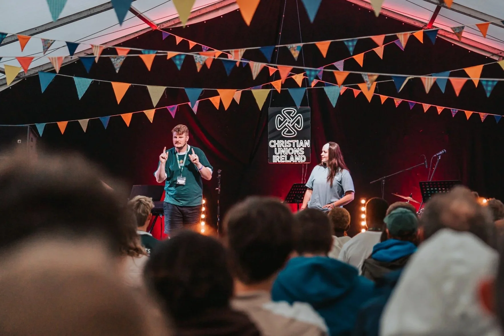 A speaker on stage at a Christian Union Ireland event, with colorful triangular banners hanging overhead and a sign behind him that reads "Christian Unions Ireland." The audience is visible in the foreground.