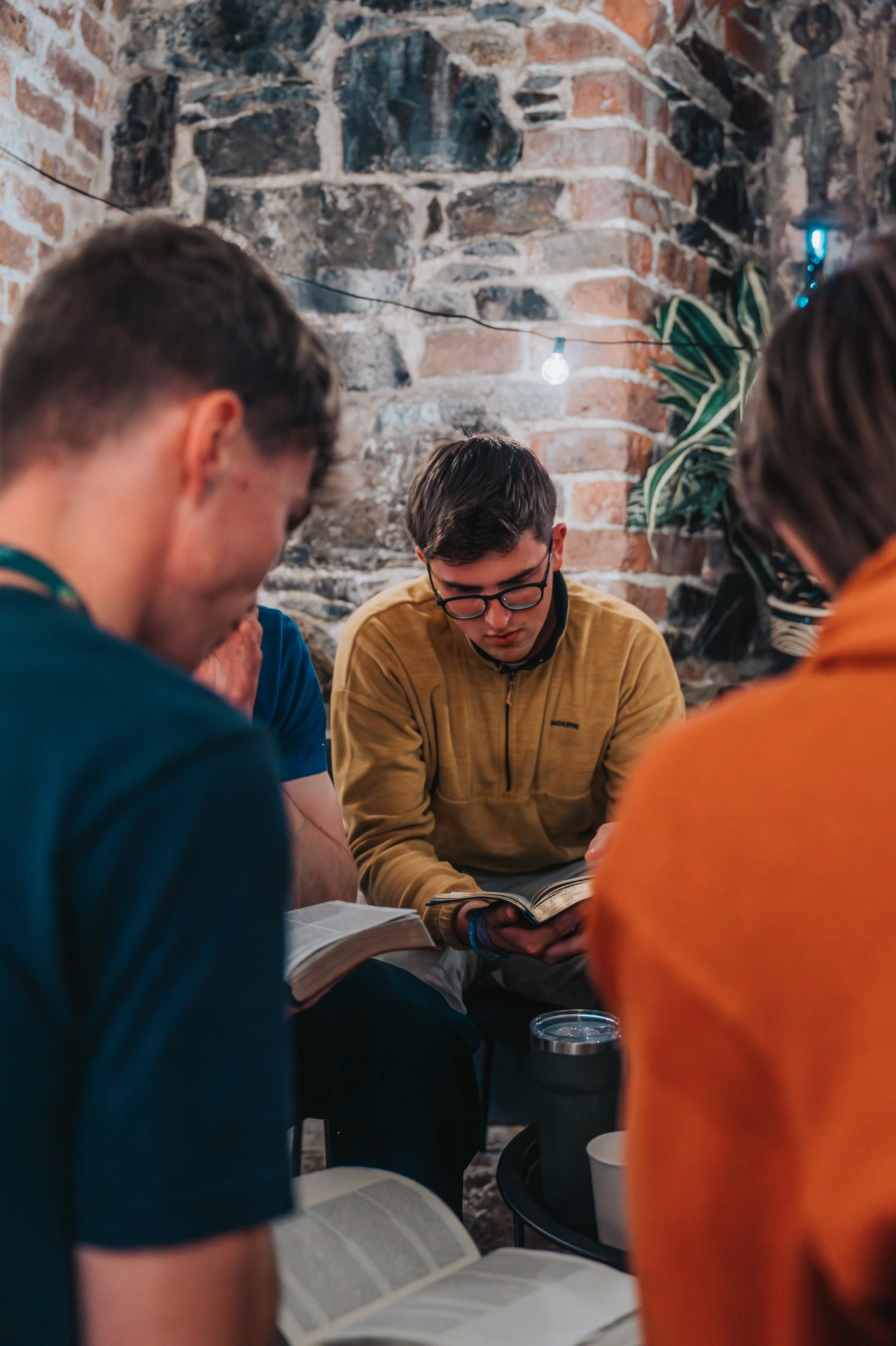 A group of young people reading books together in a cozy indoor space with an exposed brick wall and string lights. Some are wearing jackets and glasses.