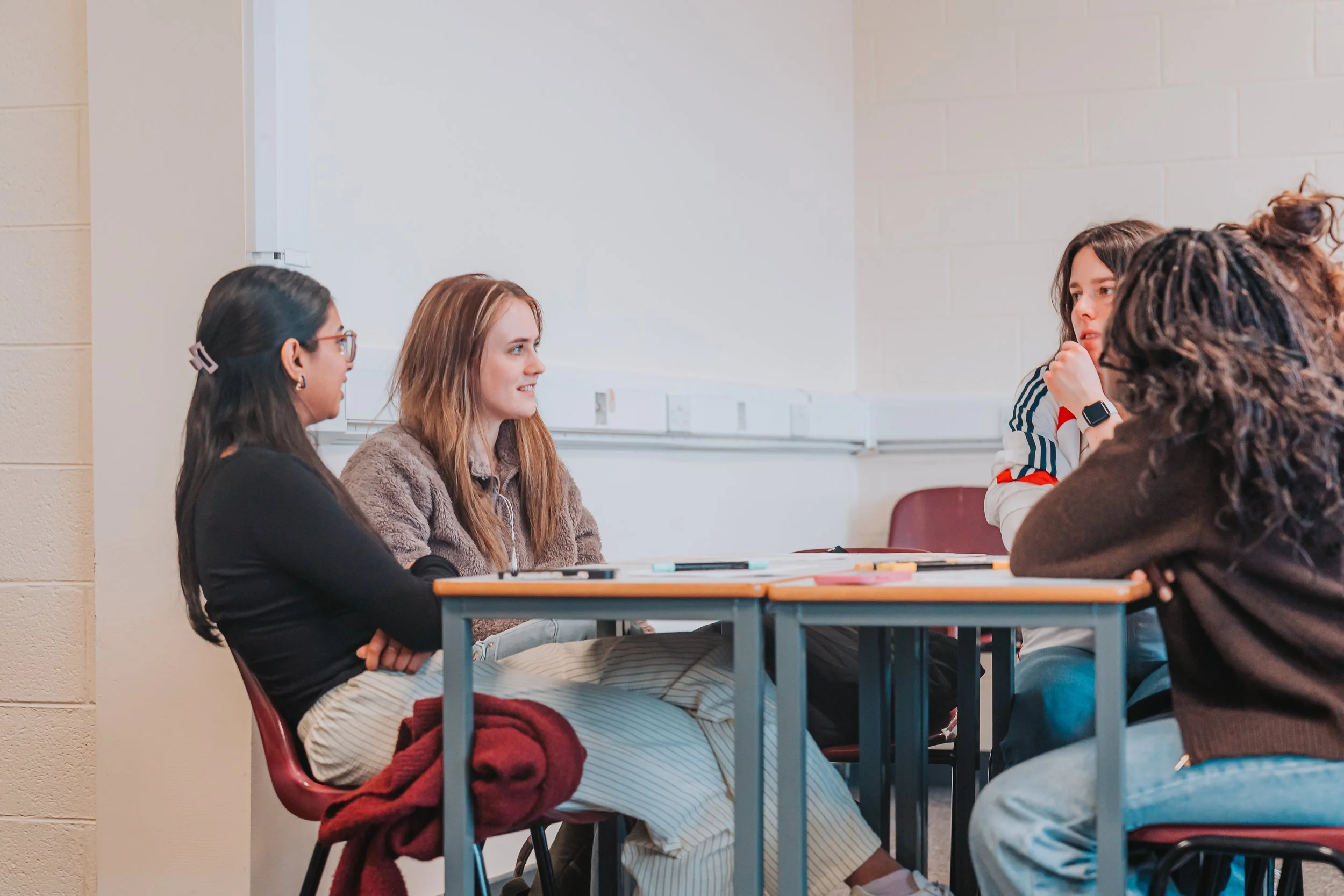 Group of four women sitting at a table engaging in conversation in a classroom or meeting room with a whiteboard in the background.