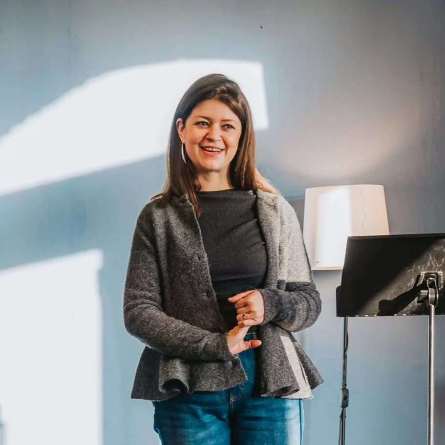 A woman with shoulder-length brown hair, smiling, standing in front of a plain wall with a lamp and a music stand nearby.