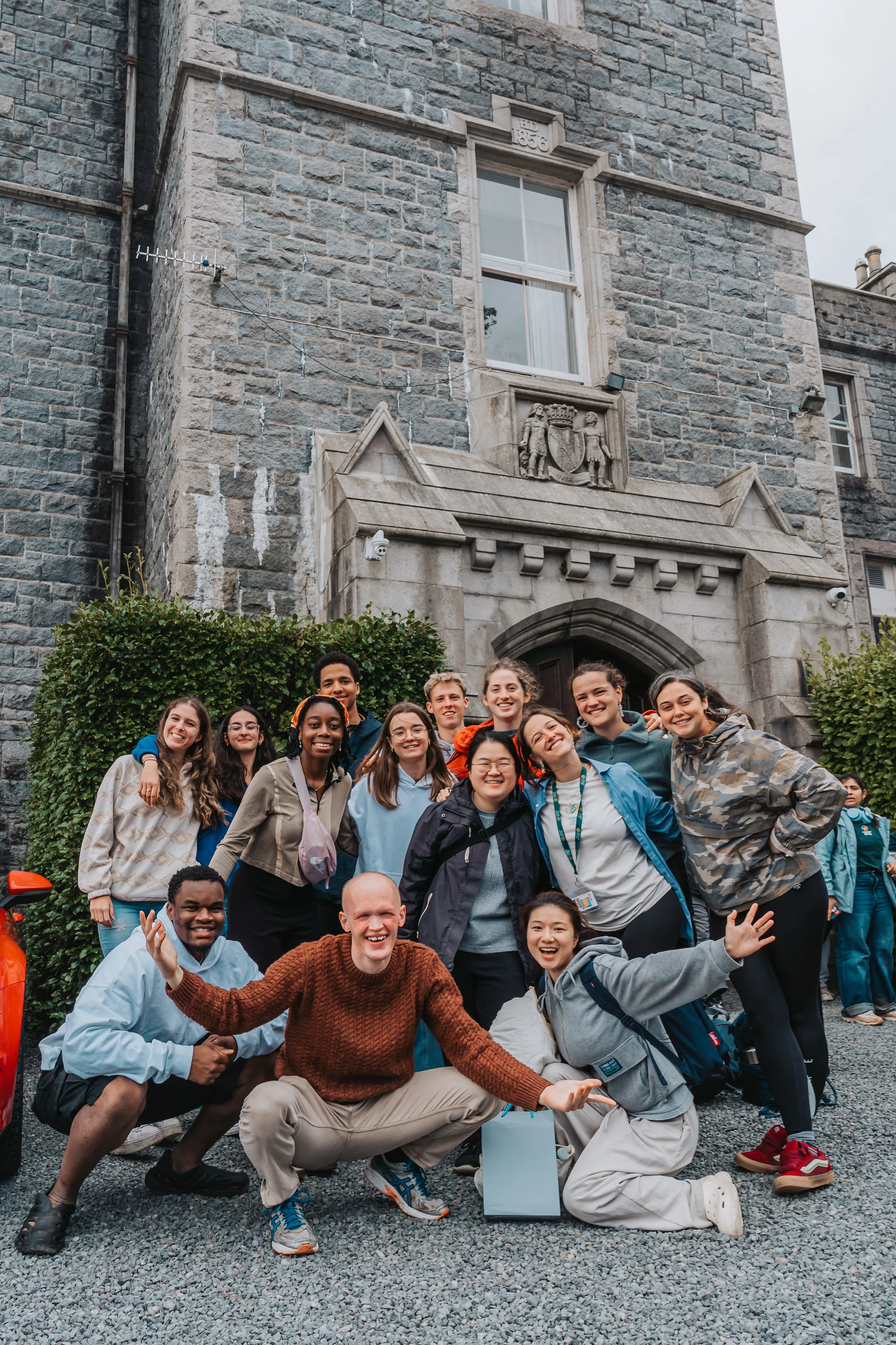 Group of smiling young people gathered outside in front of a stone building with a crest and window, posing for a photo on a gravel surface, with some crouching and others standing