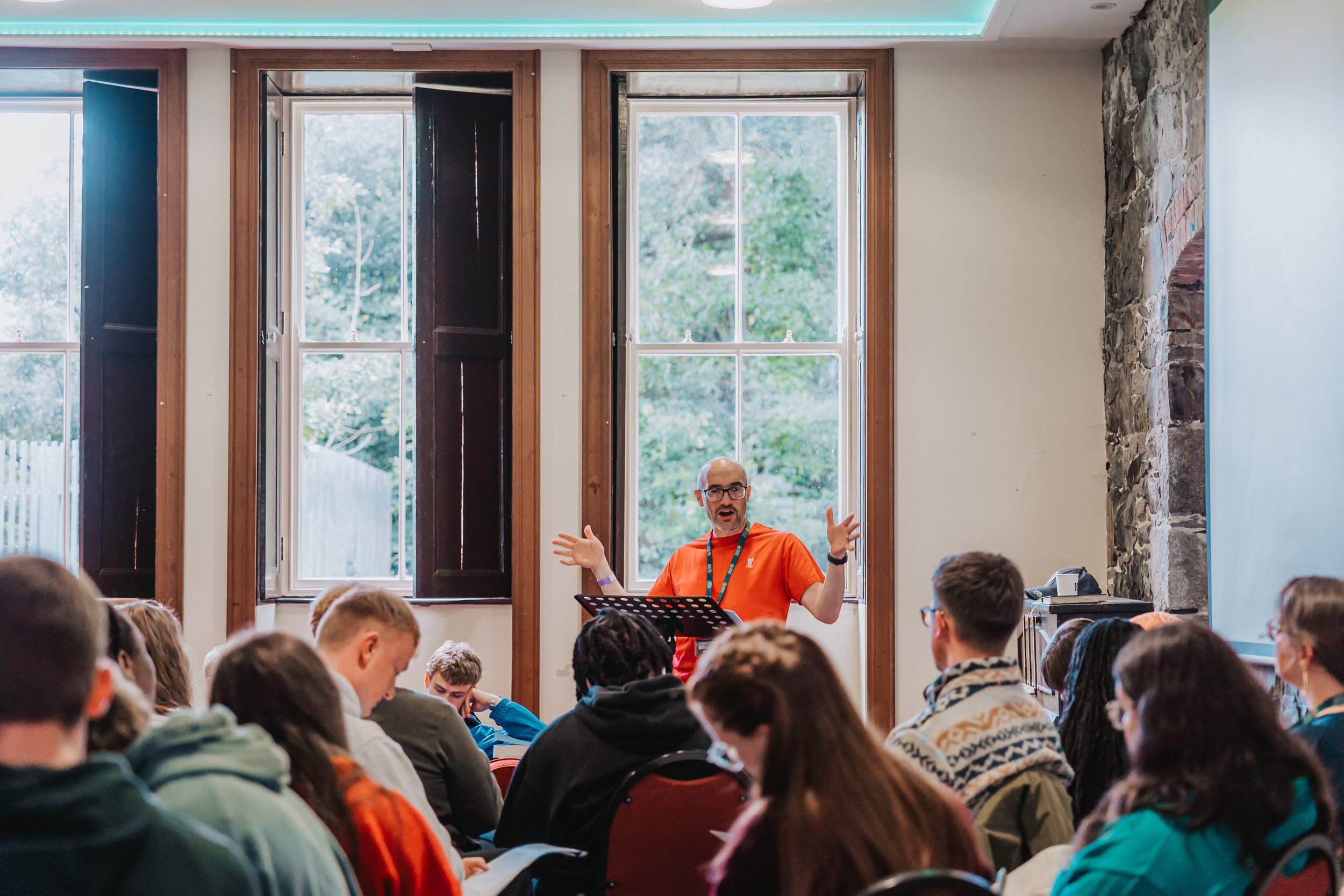 A man with glasses and a red shirt speaking or instructing in front of a seated audience inside a room with large windows, stone wall, and a blue ceiling.