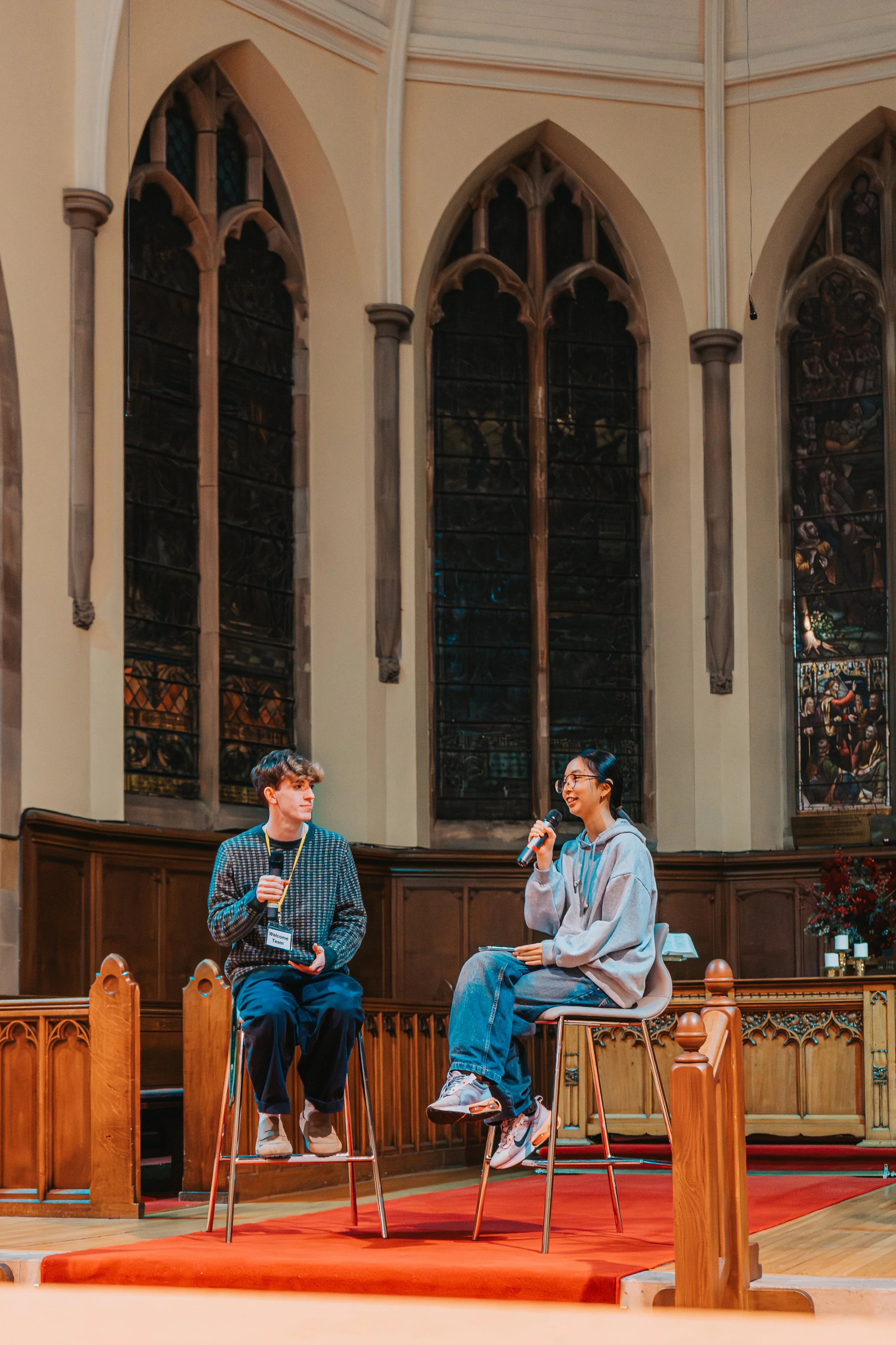 Two young people, a man and a woman, sit on stools on a stage inside a church or similar building with stained glass windows. The woman, holding a microphone and smiling, appears to be speaking, while the man, also holding a microphone, listens. They are engaged in a discussion or interview.