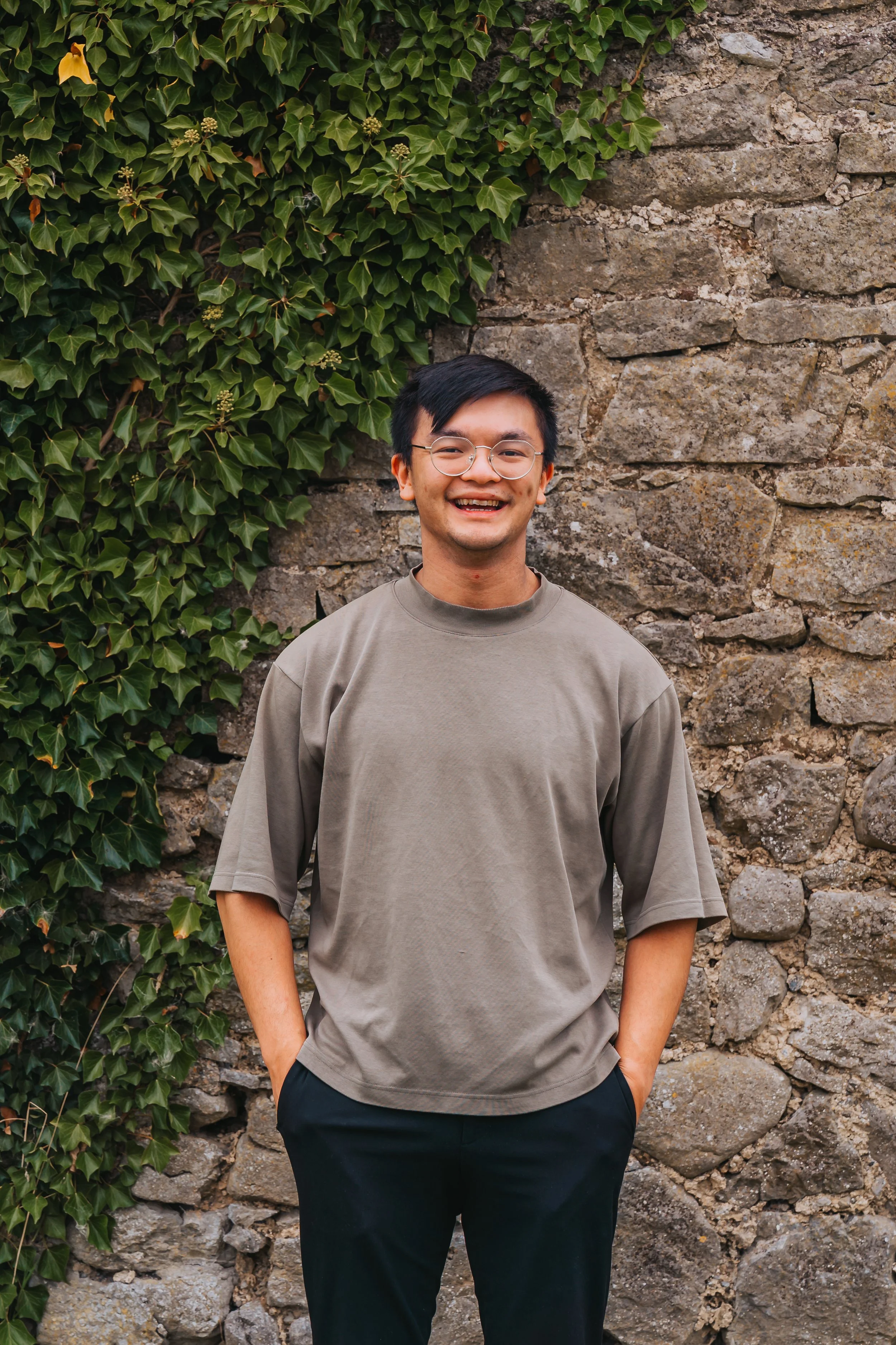 A young man with glasses, smiling, standing against a stone wall with green ivy on the left side.