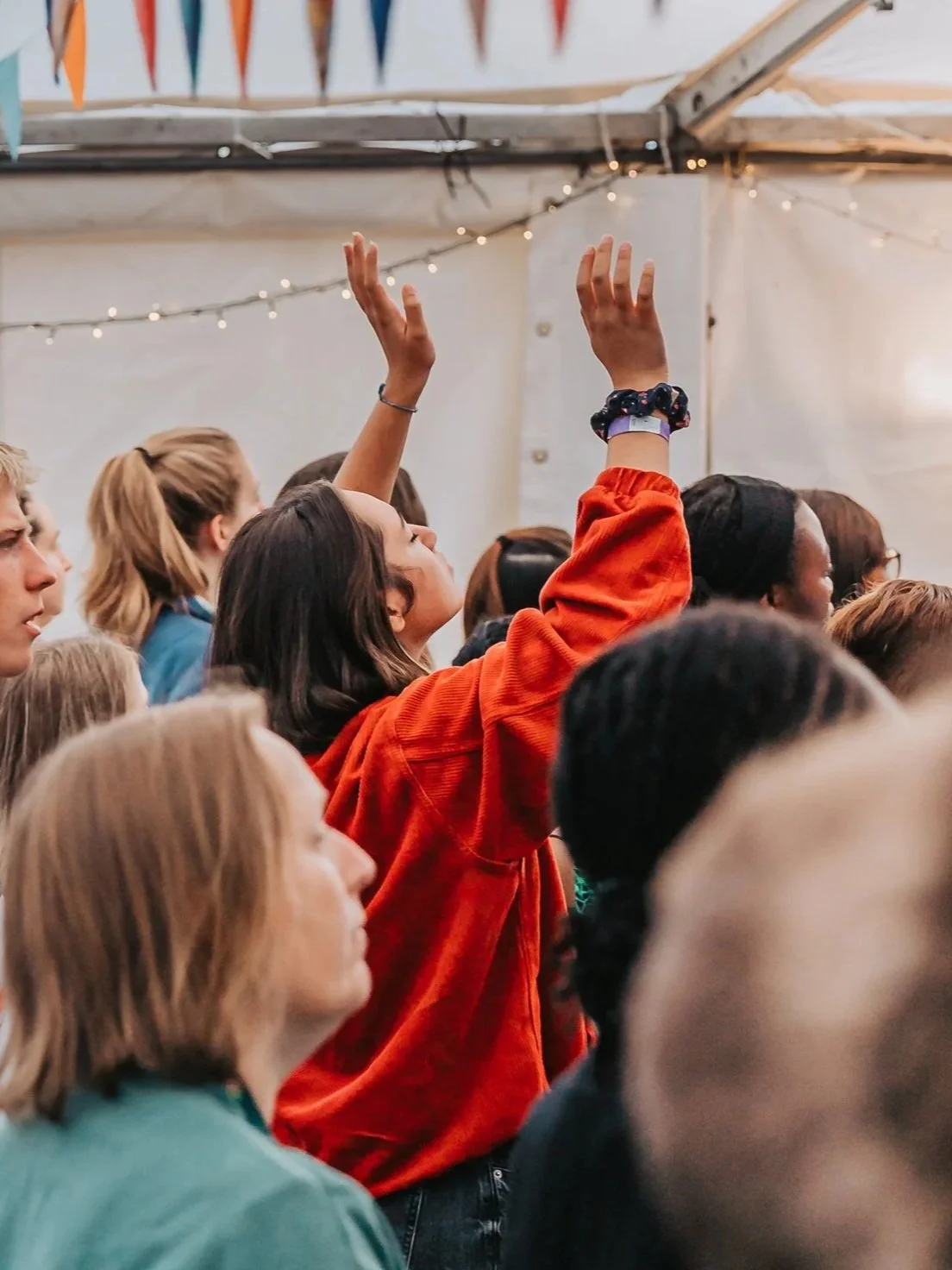 A group of people gathered indoors under a tent-like structure, with some individuals raising their hands.