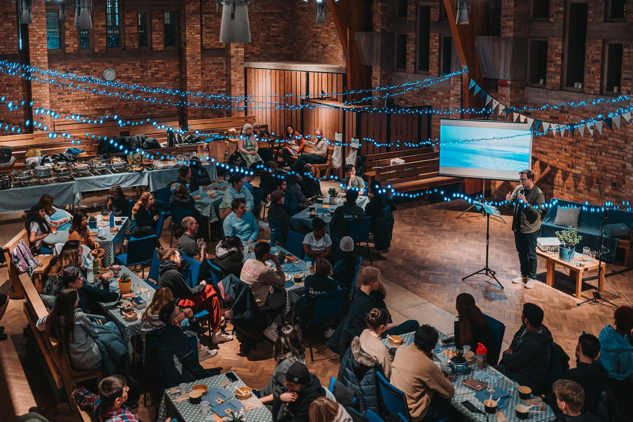 People seated at decorated tables in a brick hall during an event, with a speaker giving a presentation on a screen and hanging blue string lights and pennant banners overhead.