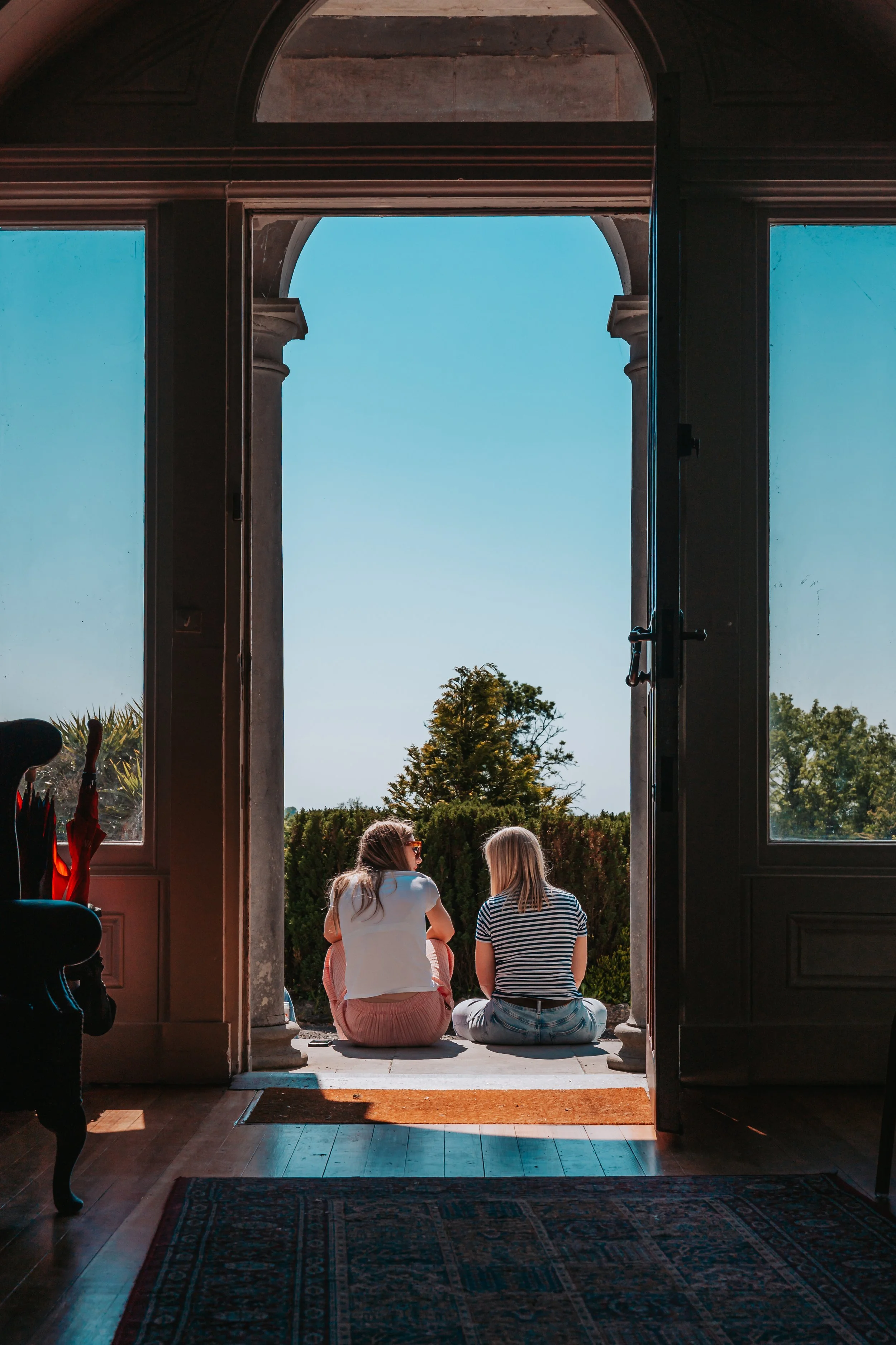 Two women sitting on the ground in front of an open archway, looking outside at clear blue sky and green trees.