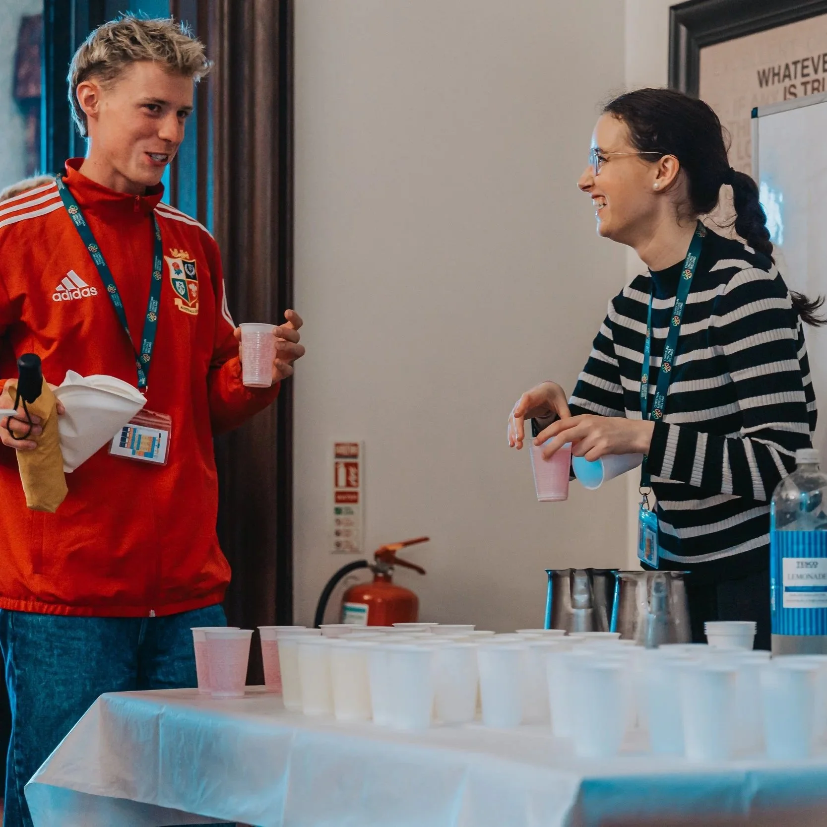 Two women smiling and talking at a table with cups and metal shakers, in an indoor setting with a fire extinguisher and framed poster on the wall.