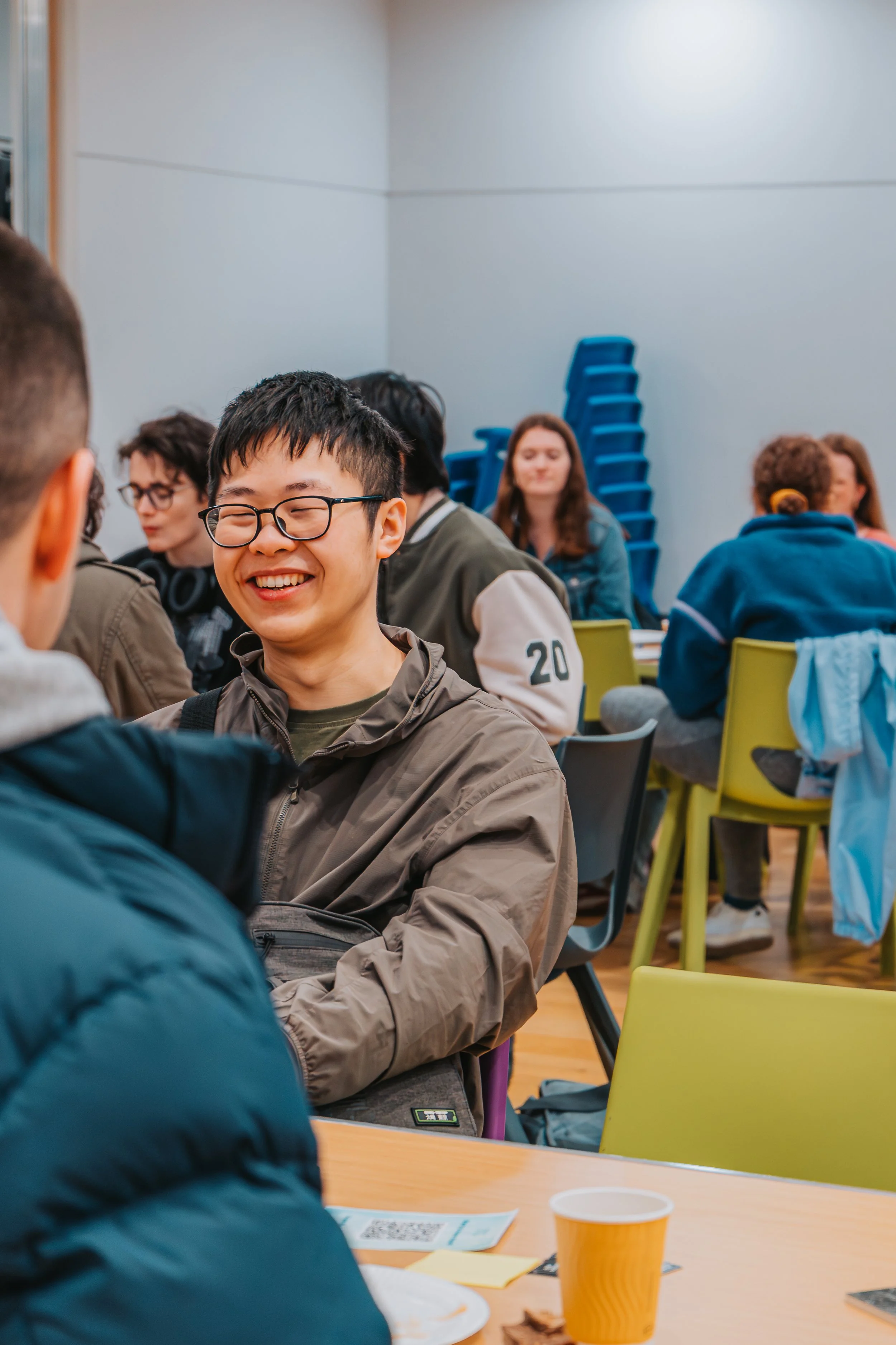 A group of young people sitting around a table in a room, with a stack of blue chairs against the wall, smiling and engaging in conversation.