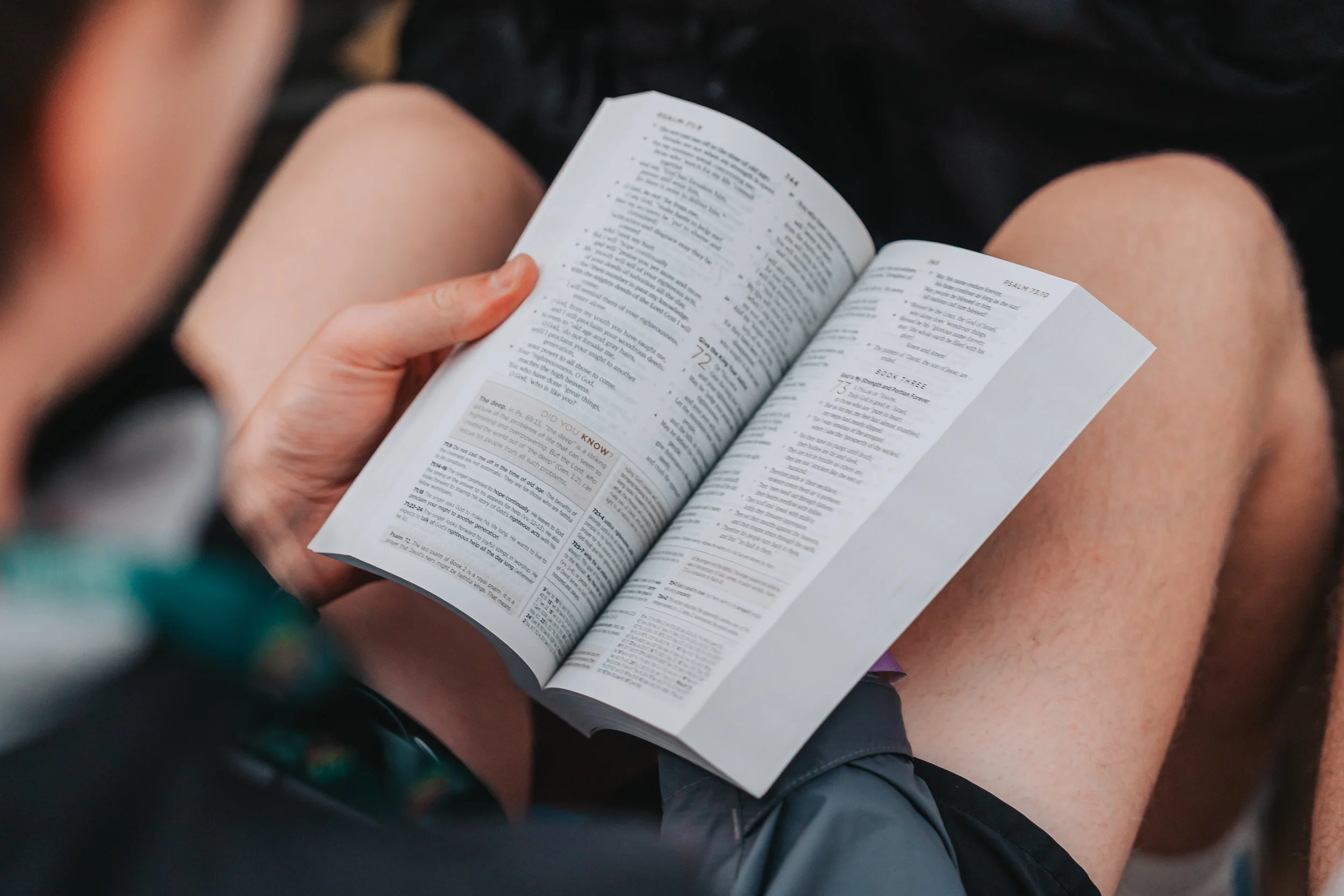 Person holding open paperback Bible sitting with legs crossed, wearing shorts.