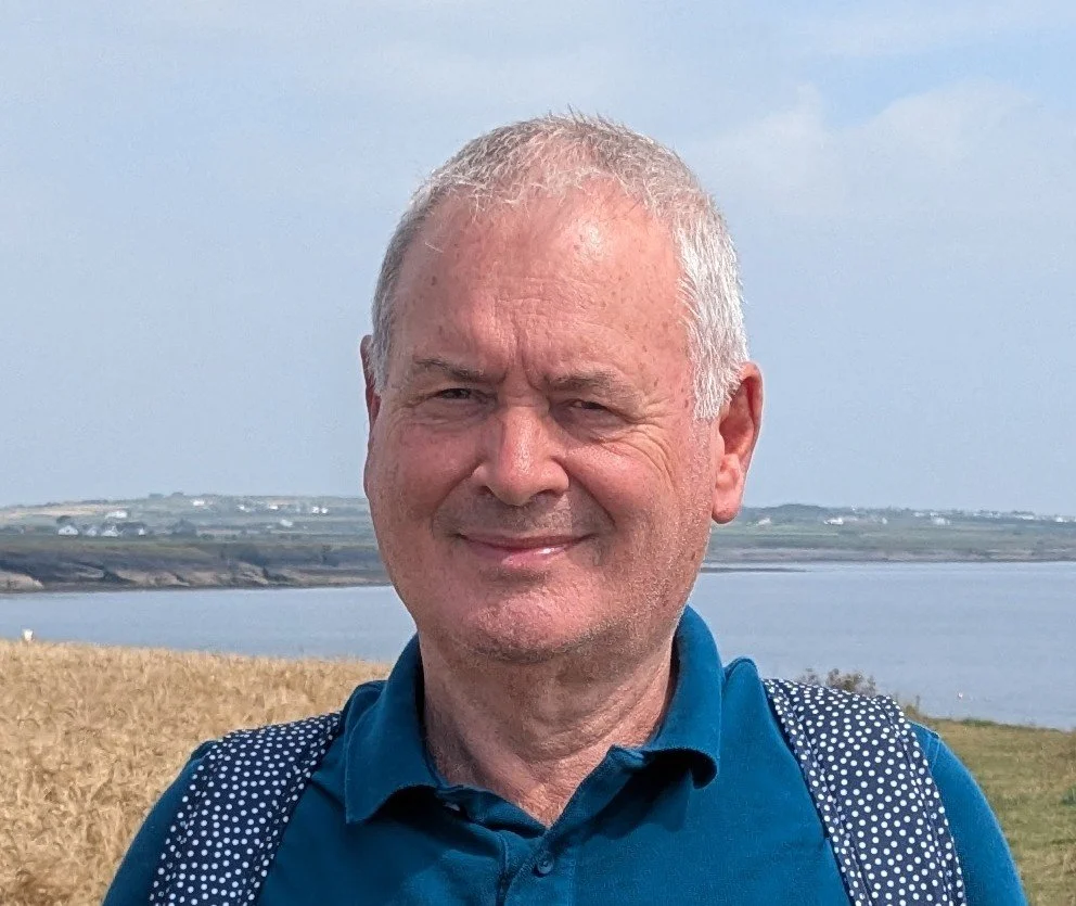 A smiling middle-aged man with short gray hair, wearing a blue shirt and backpack, standing outdoors near a body of water with fields in the background.