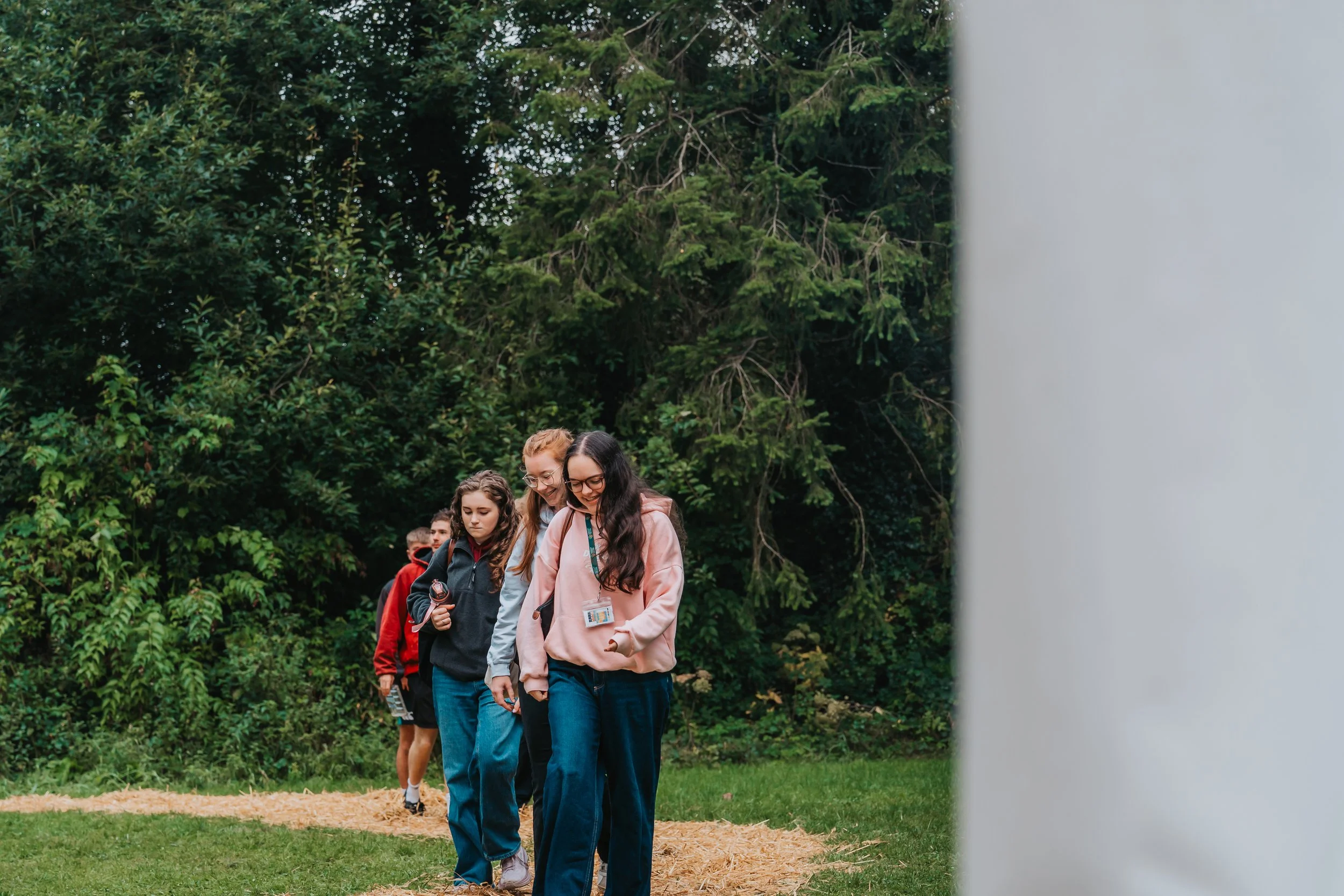 A group of young people walking in a line outdoors on a trail surrounded by green trees, with a partial view of a white structure on the right side.