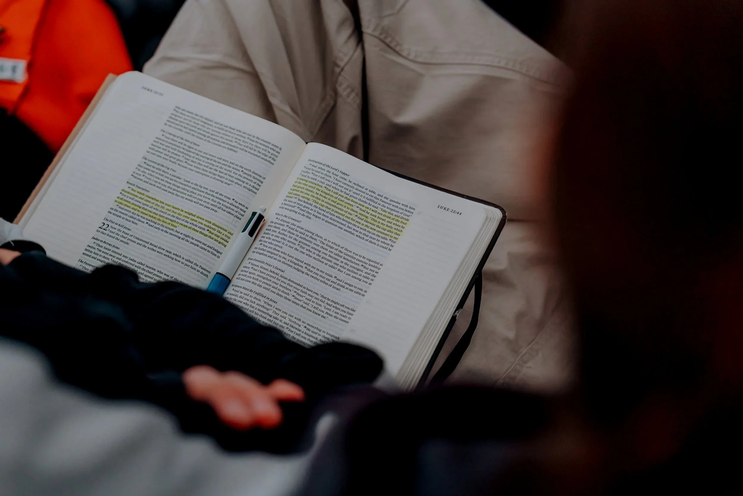 Person sitting on the ground reading a Bible, with a focus on the open book and highlights on the pages, and a backpack next to them.