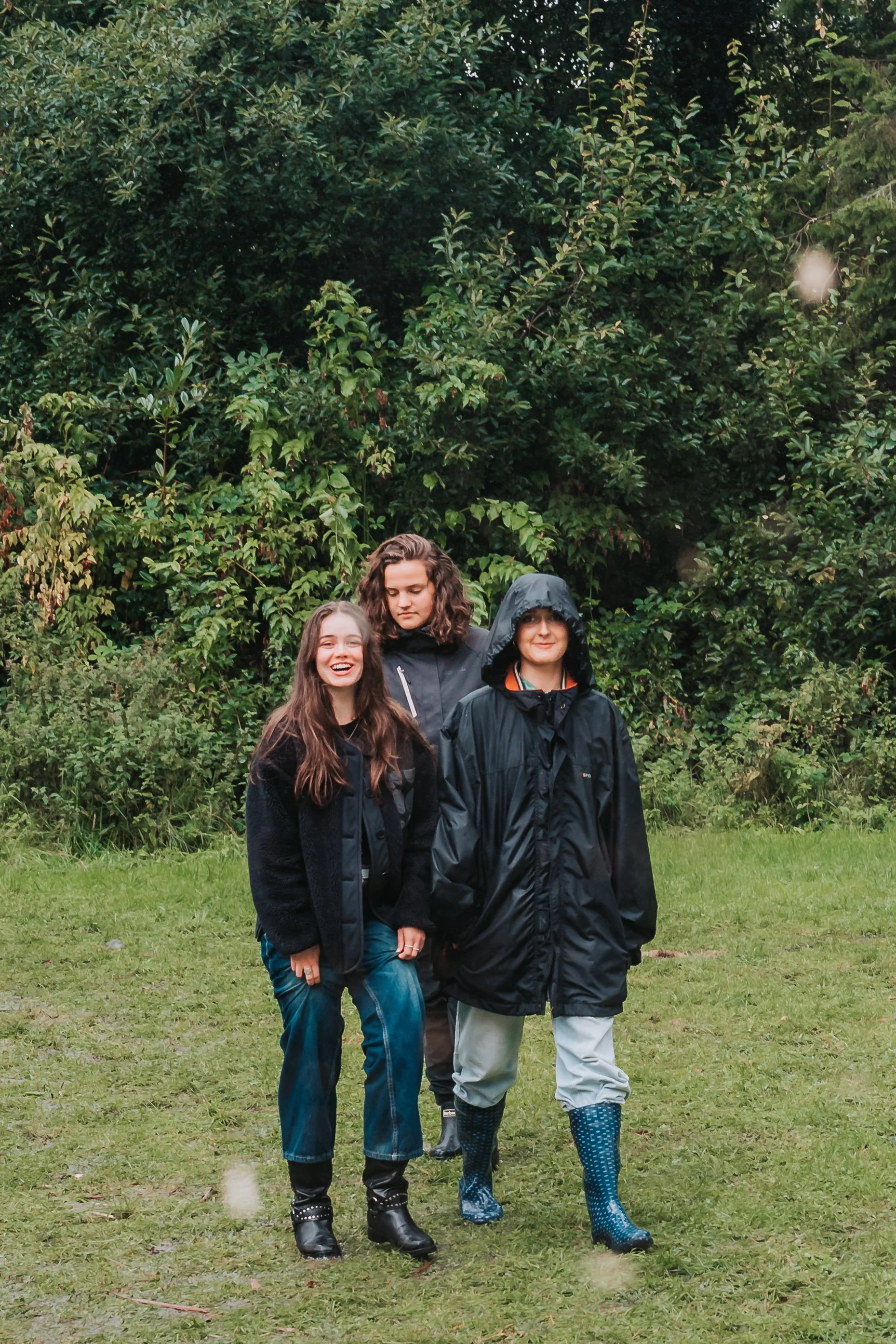 Three friends walking outdoors on a grassy area in front of lush green trees. Two women and one man are dressed in rain jackets and waterproof boots, smiling and enjoying the day.