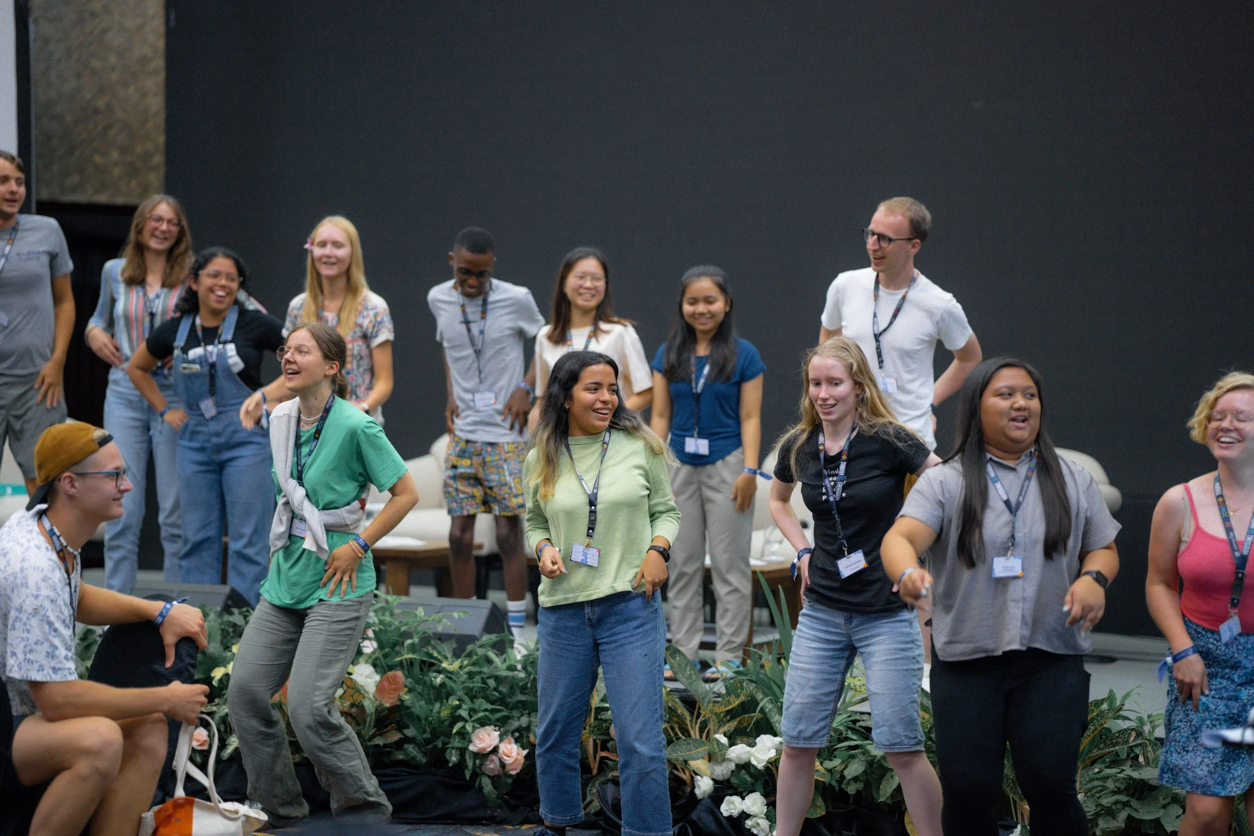 A group of diverse people, mostly young women, are standing and laughing in a room with dark walls. Some are wearing conference badges. There are plants and flowers in the foreground.