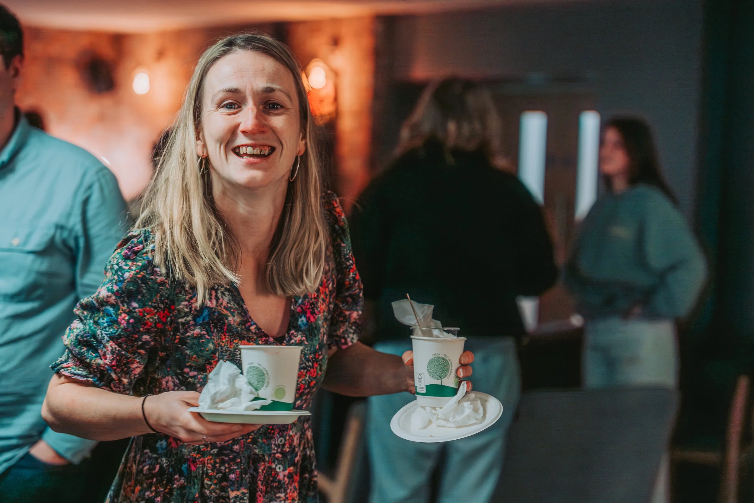 A woman with long blonde hair and a floral dress is holding a tray with two cups of coffee and napkins at a social gathering or event. She is smiling and appears to be enjoying the moment. There are blurred people in the background.
