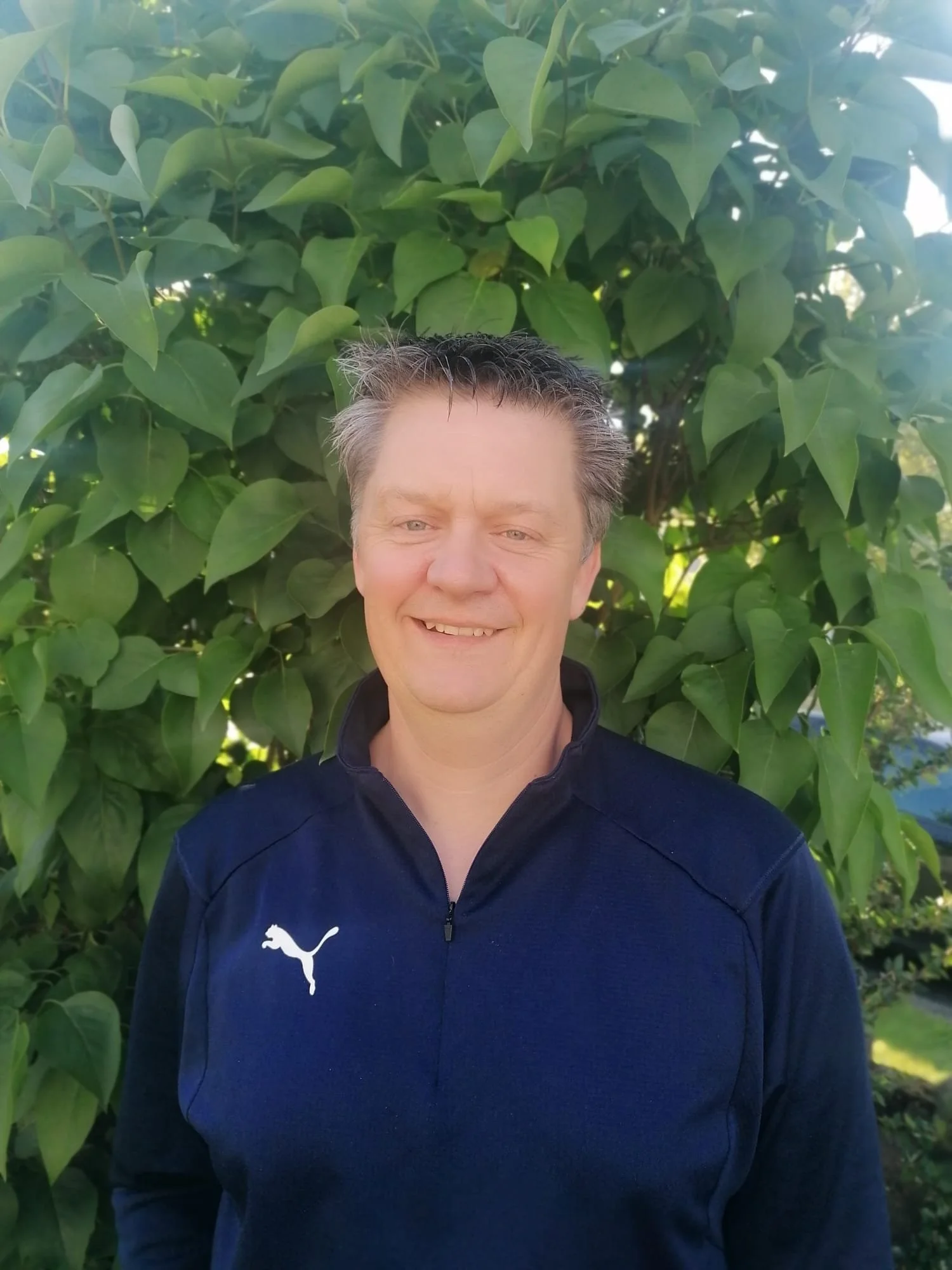 A smiling man with short gray hair wearing a dark blue Puma sports jacket standing in front of green leafy bushes.