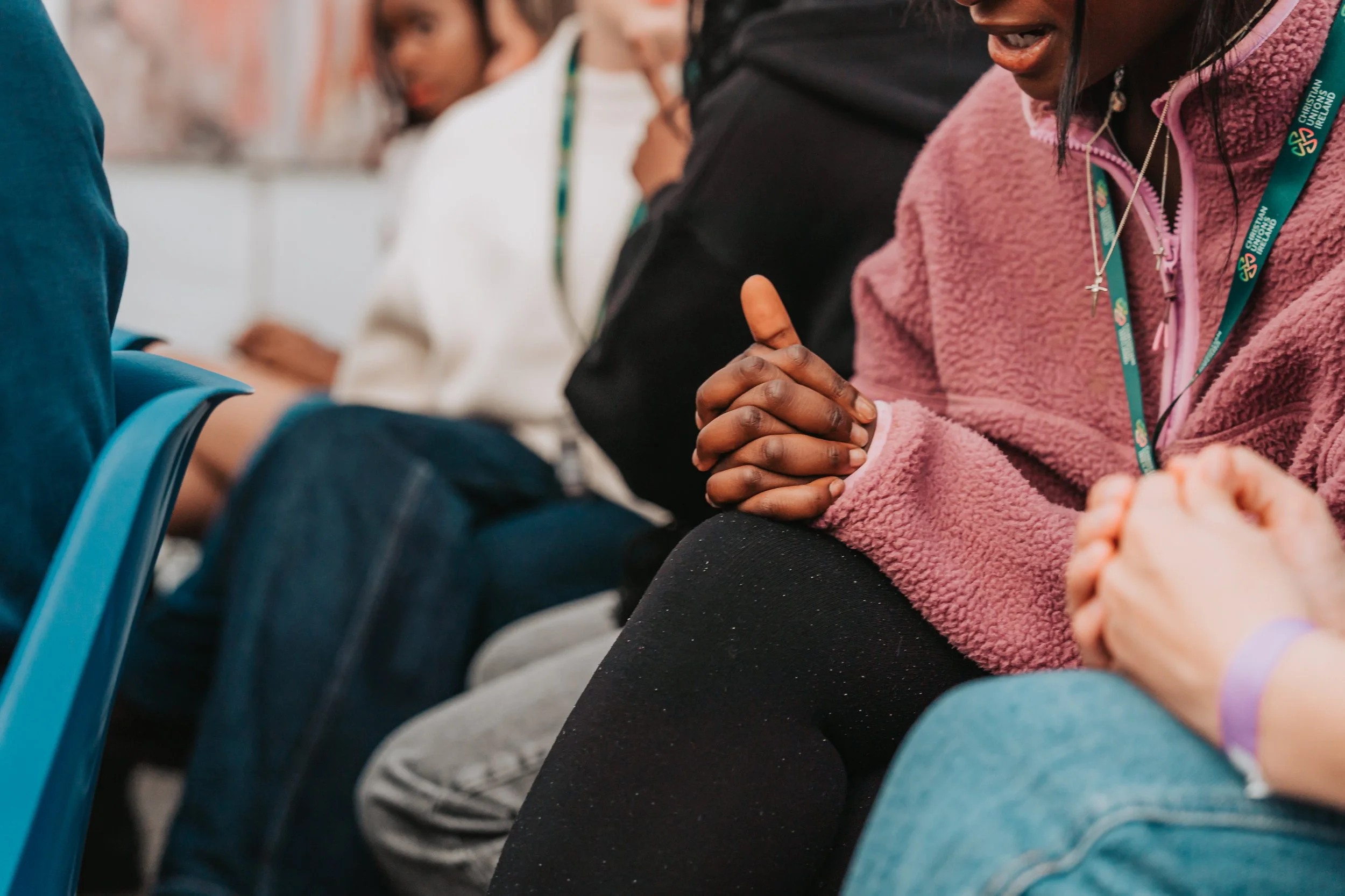 A woman wearing a pink fleece jacket and a black necklace with a cross pendant, praying with her hands clasped.