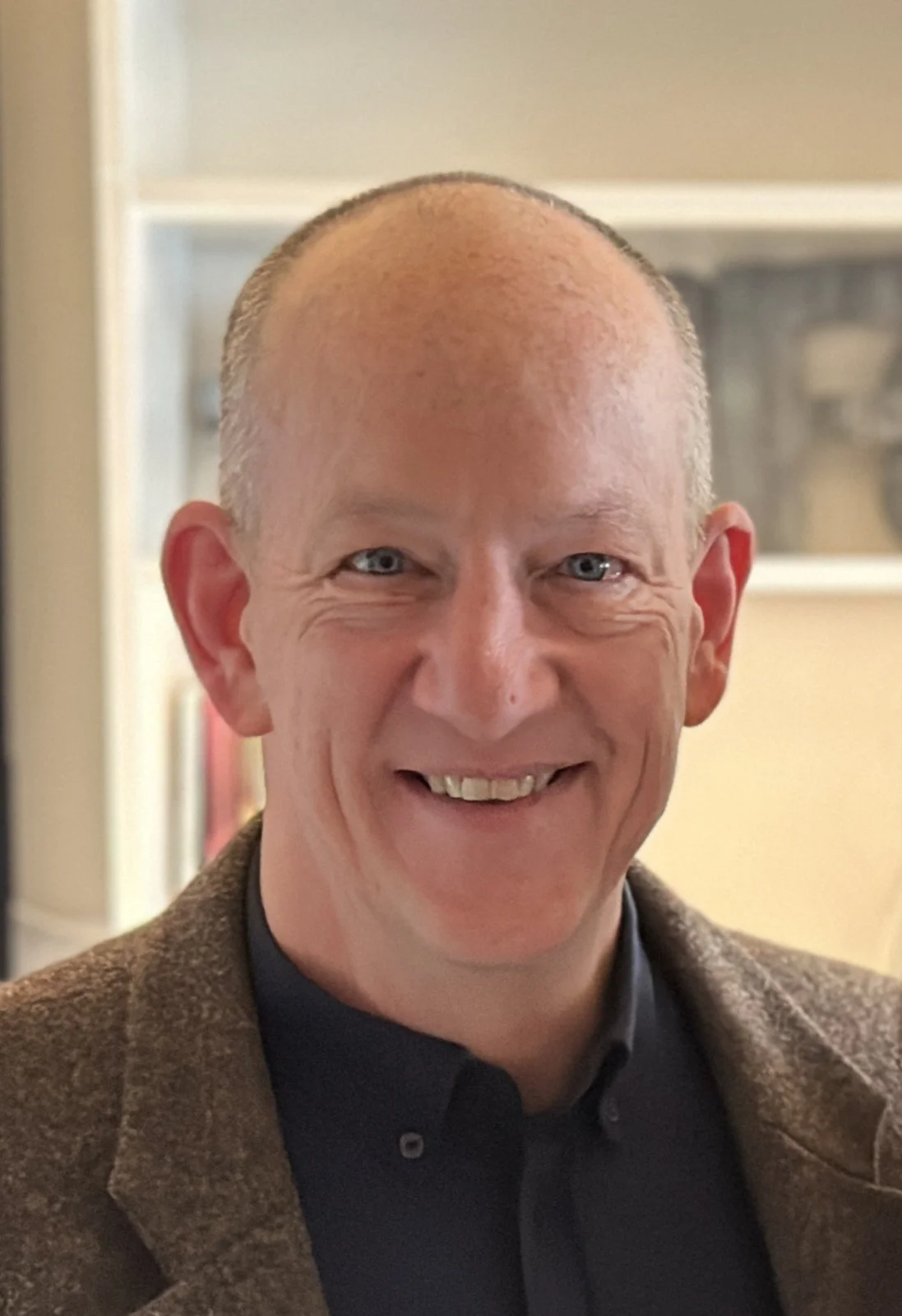 A smiling man with short hair, wearing a black shirt and a brown blazer, indoors with bookshelves and decorative items in the background.
