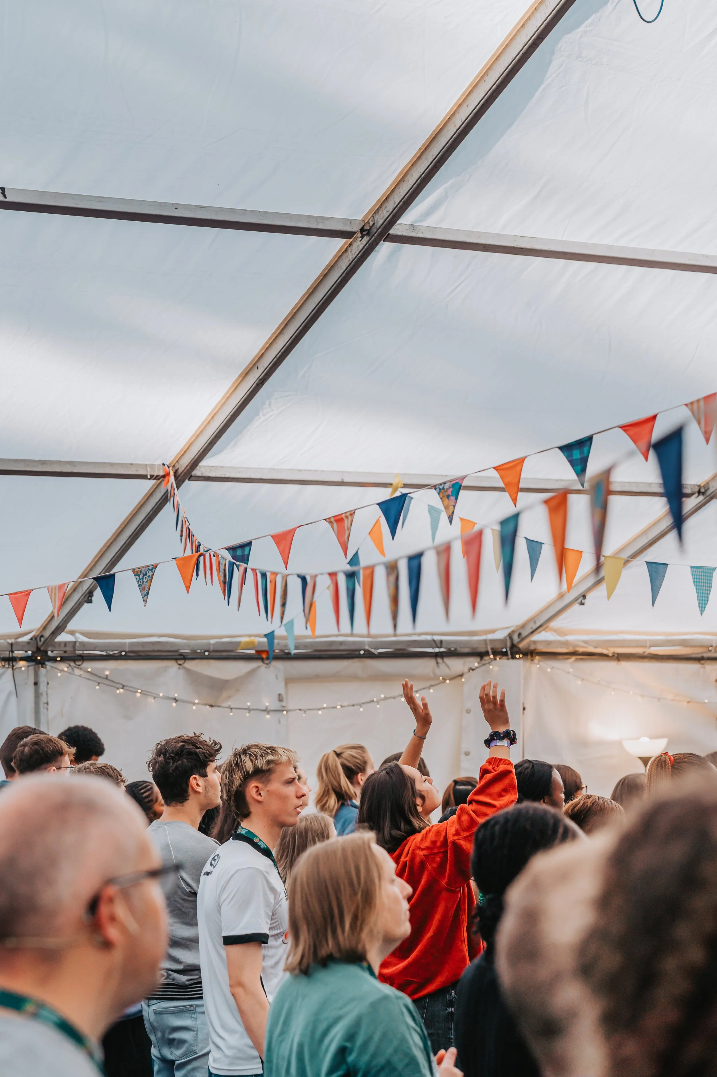 A diverse group of people participating in an indoor event under a tent decorated with colorful pennant banners, with some individuals raising their hands.