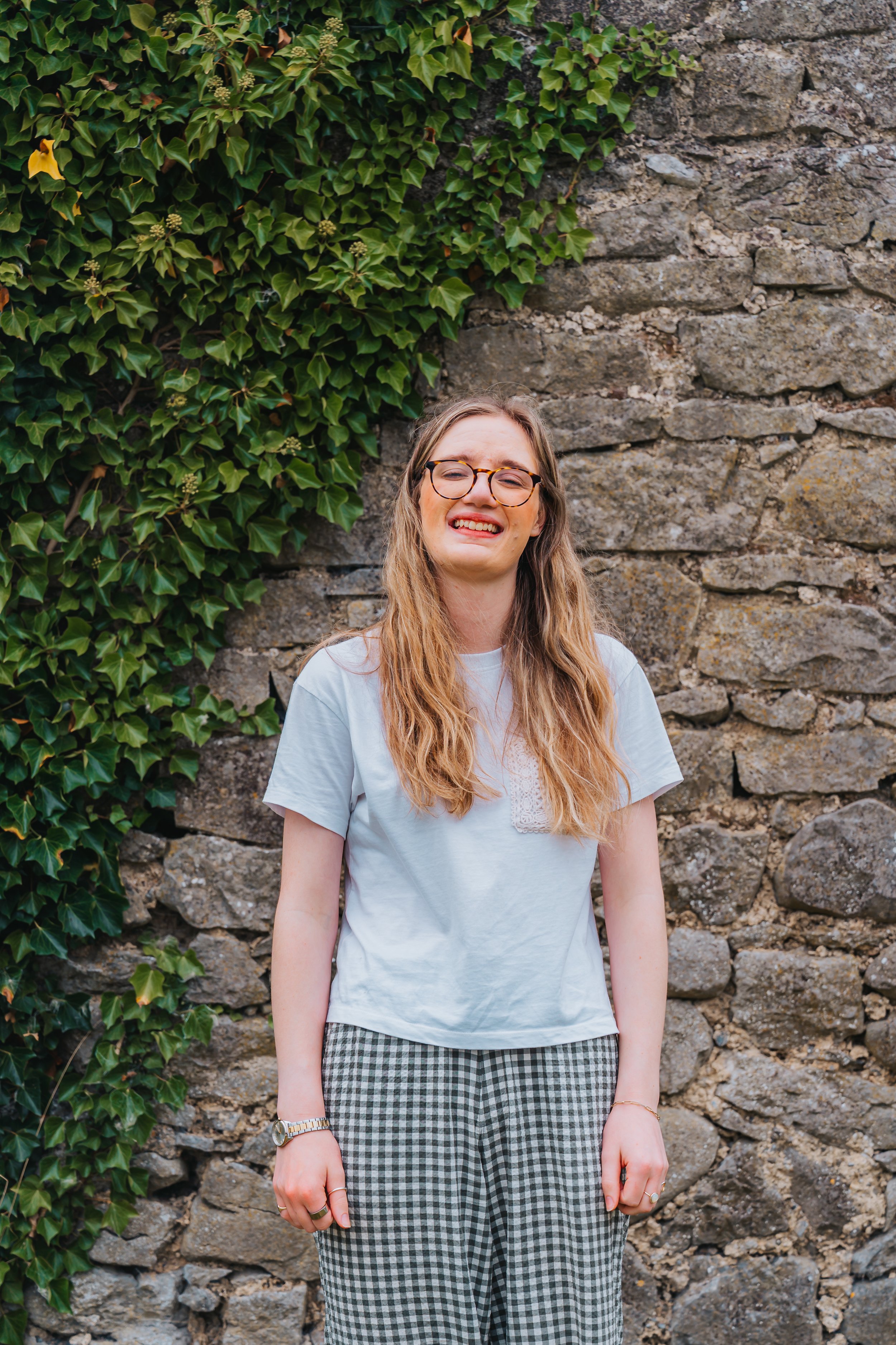A young woman with long wavy hair, glasses, and a wide smile standing outdoors against a stone wall with green ivy plants growing on it.