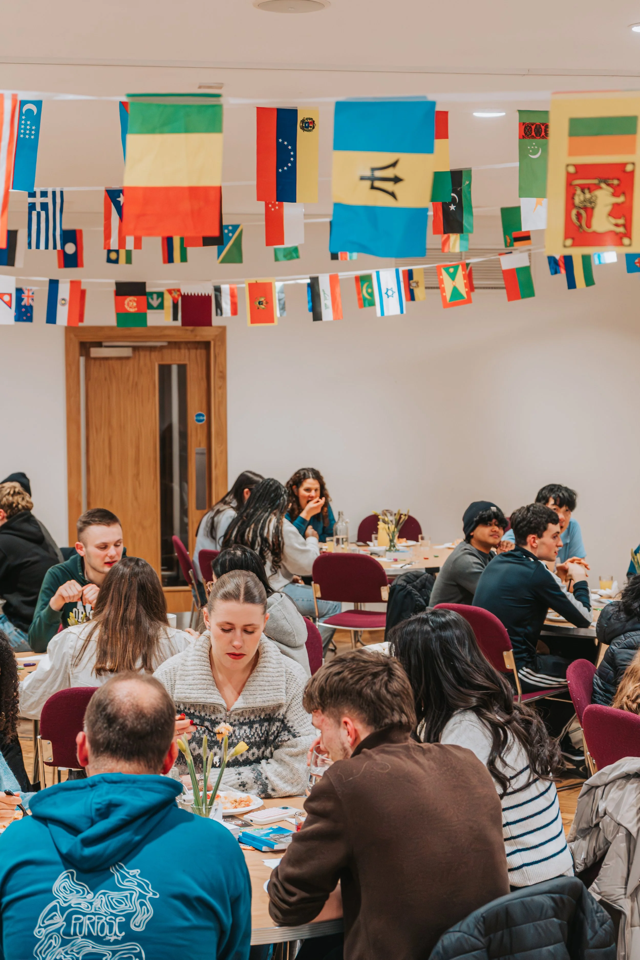 People sitting around tables at a gathering or event, with flags hanging from the ceiling.