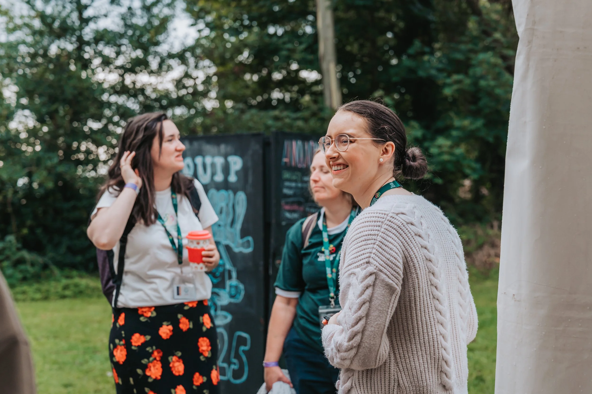Group of women outdoors, talking and smiling, with greenery in the background.