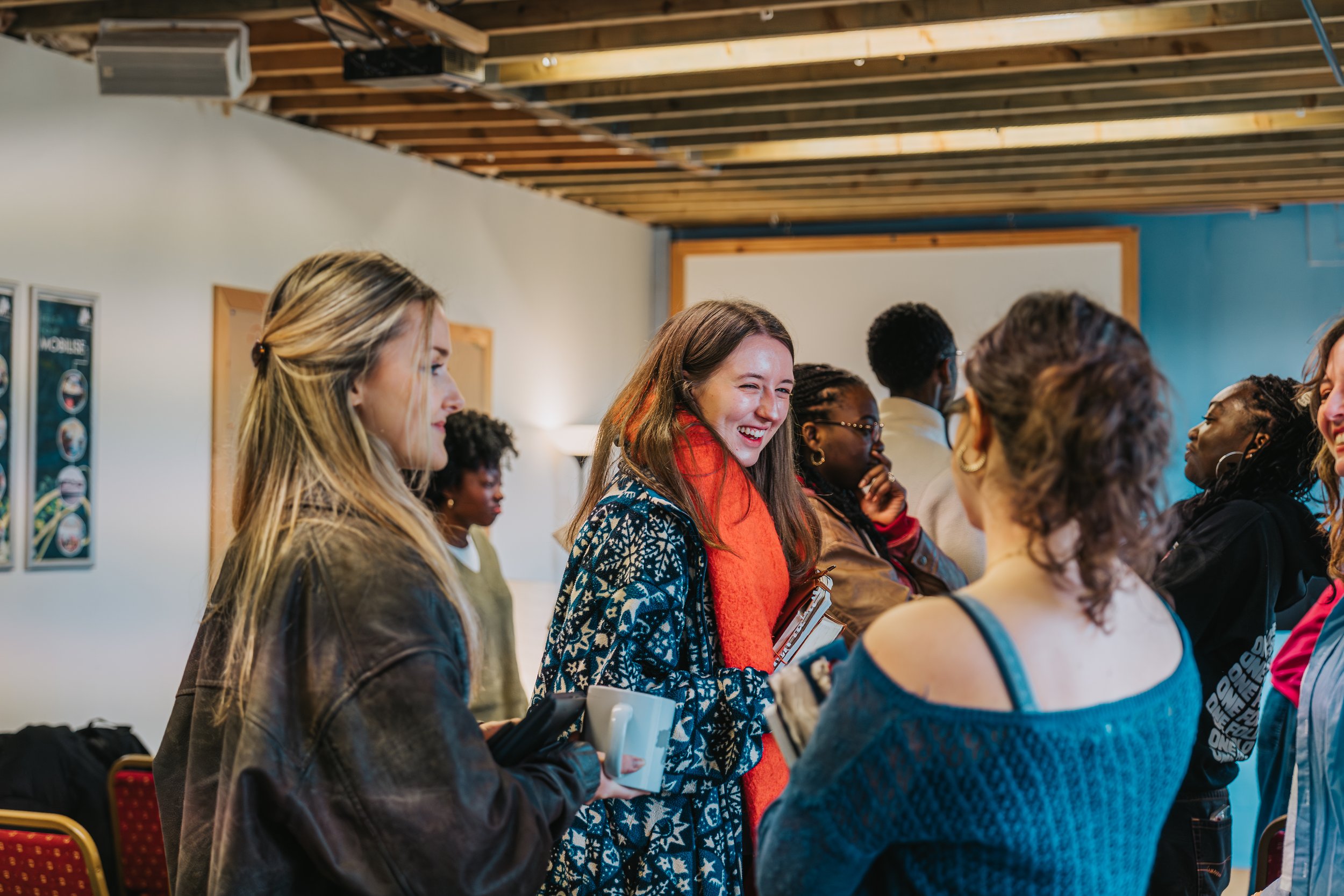 Group of diverse women gathered indoors, engaging and smiling during a social or community event, with chairs and posters visible in the background.