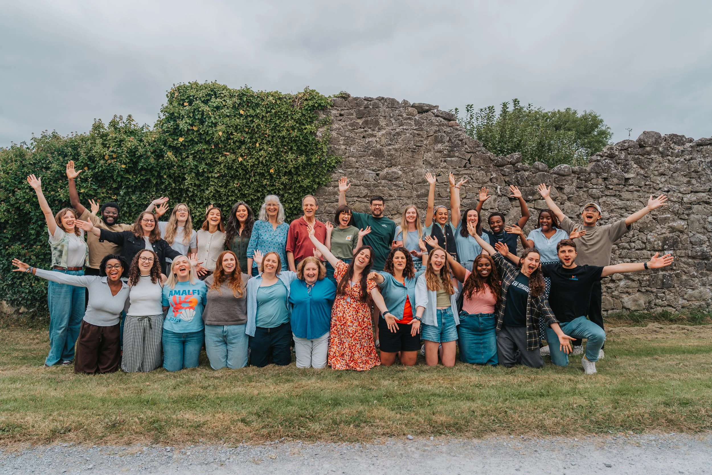 A diverse group of smiling people posing together outdoors in front of a stone wall with greenery, with some raising their hands and showing excitement.
