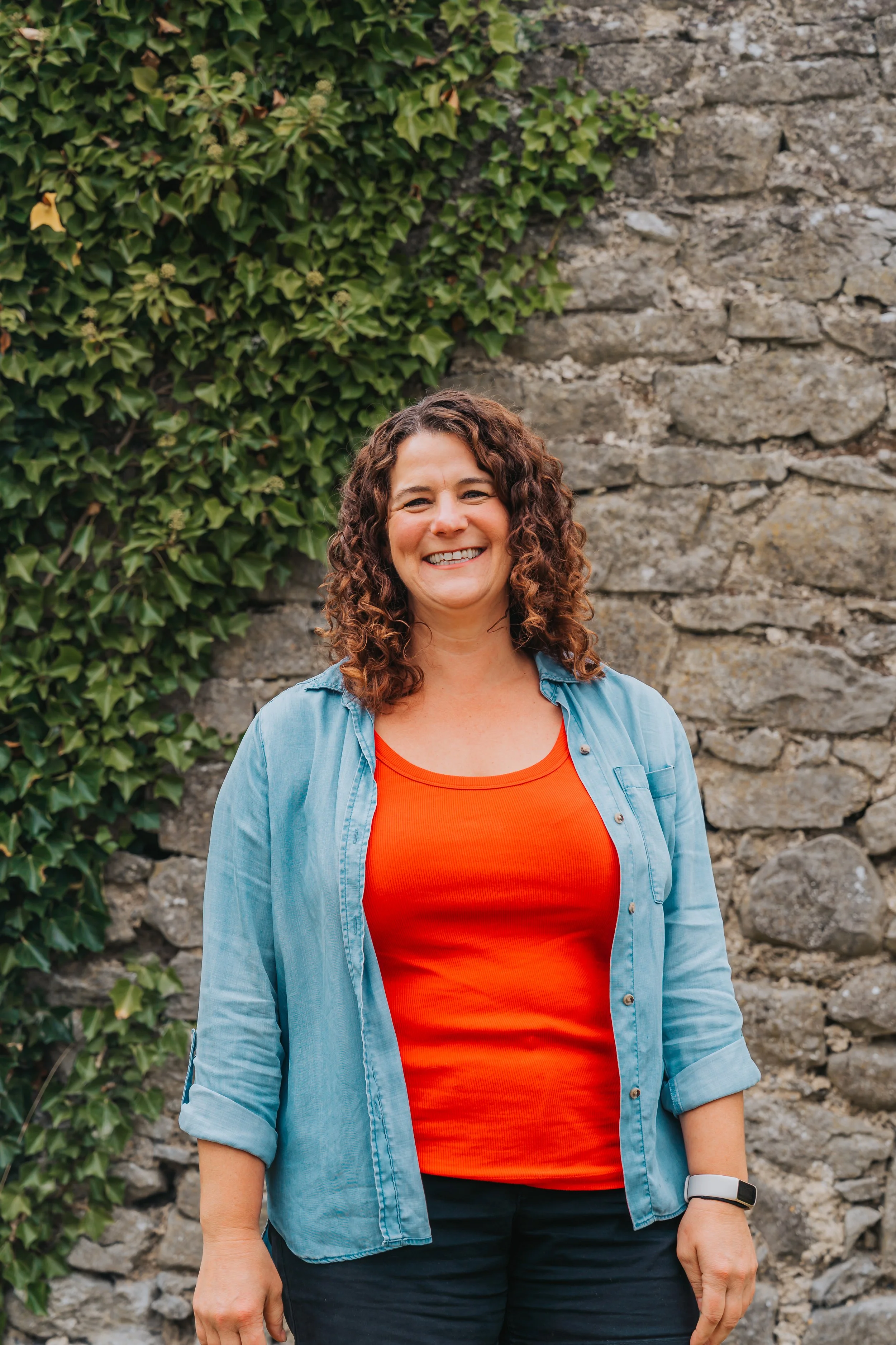 A woman with curly brown hair smiling, wearing a red top with an orange shirt underneath and a light denim button-up shirt over it, standing in front of a stone wall with ivy.