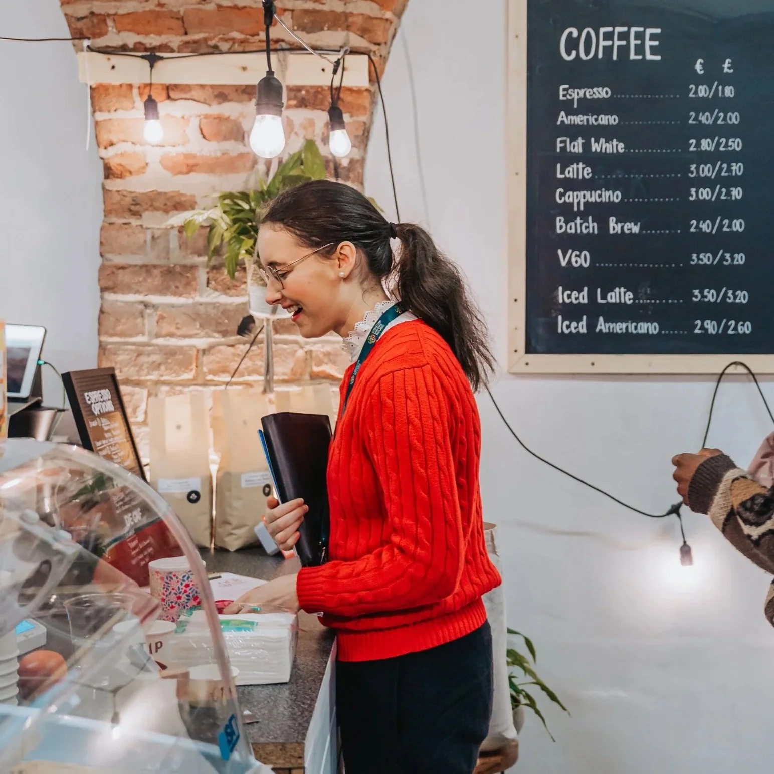 A young woman with glasses and dark hair tied in a ponytail, wearing a red sweater and a lanyard, smiling and standing at a coffee shop counter with a menu board in the background. The menu lists various types of coffee and prices.