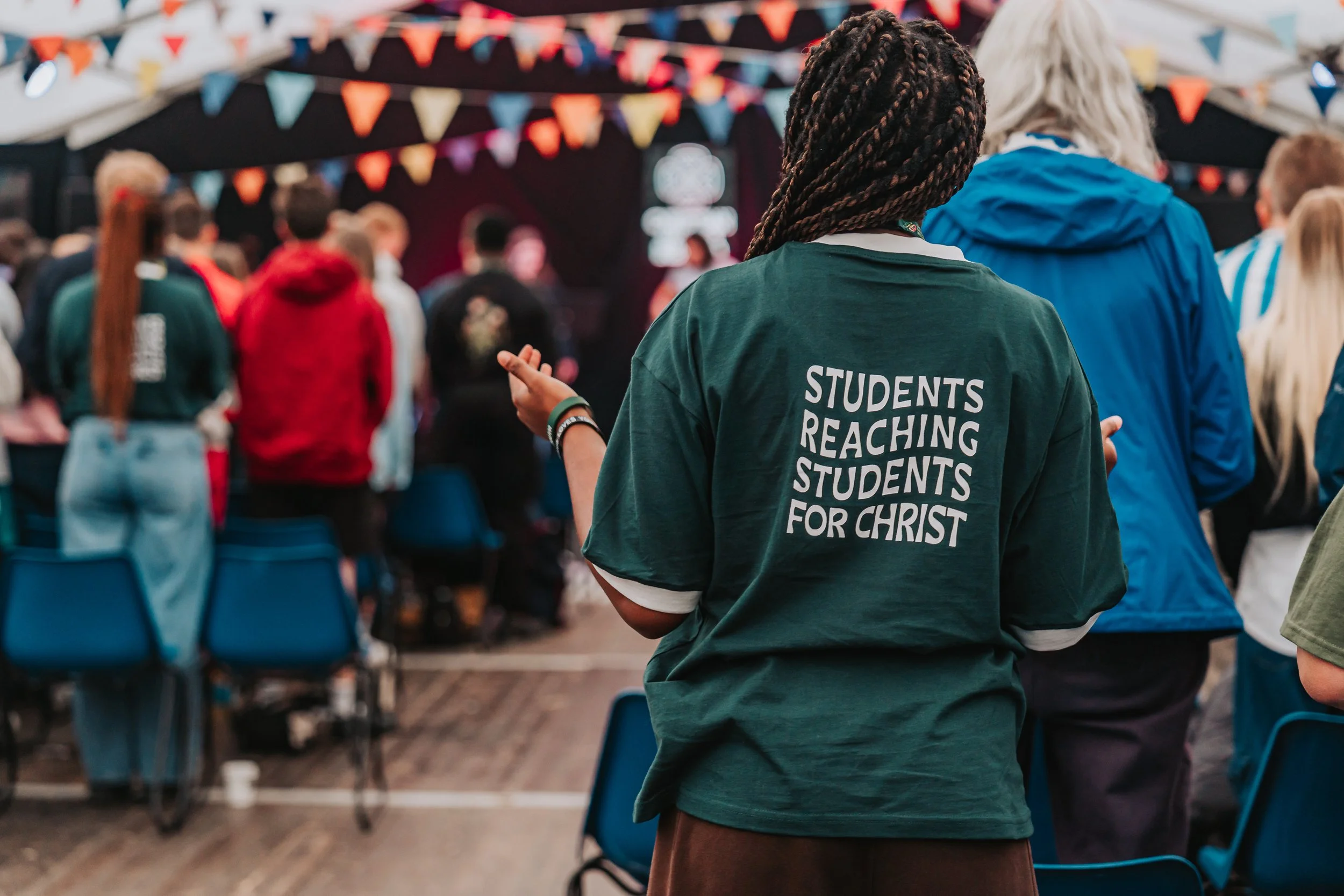 A person wearing a green hoodie with the words 'Students reaching students for Christ' on the back, standing among a crowd at an indoor event with colorful flags and a stage in the background.