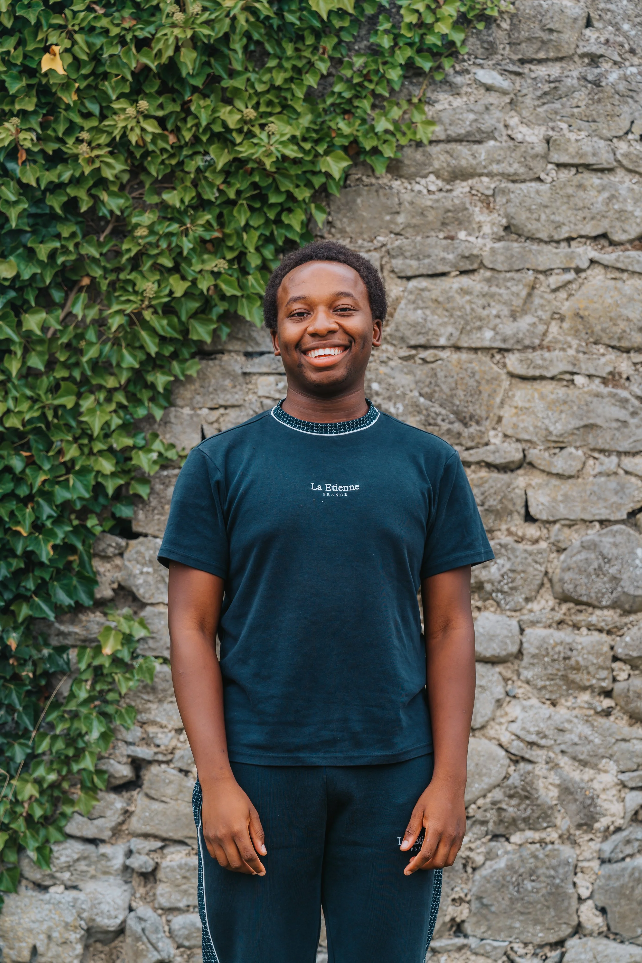 Smiling young man standing outdoors in front of a stone wall with green ivy on the left side.