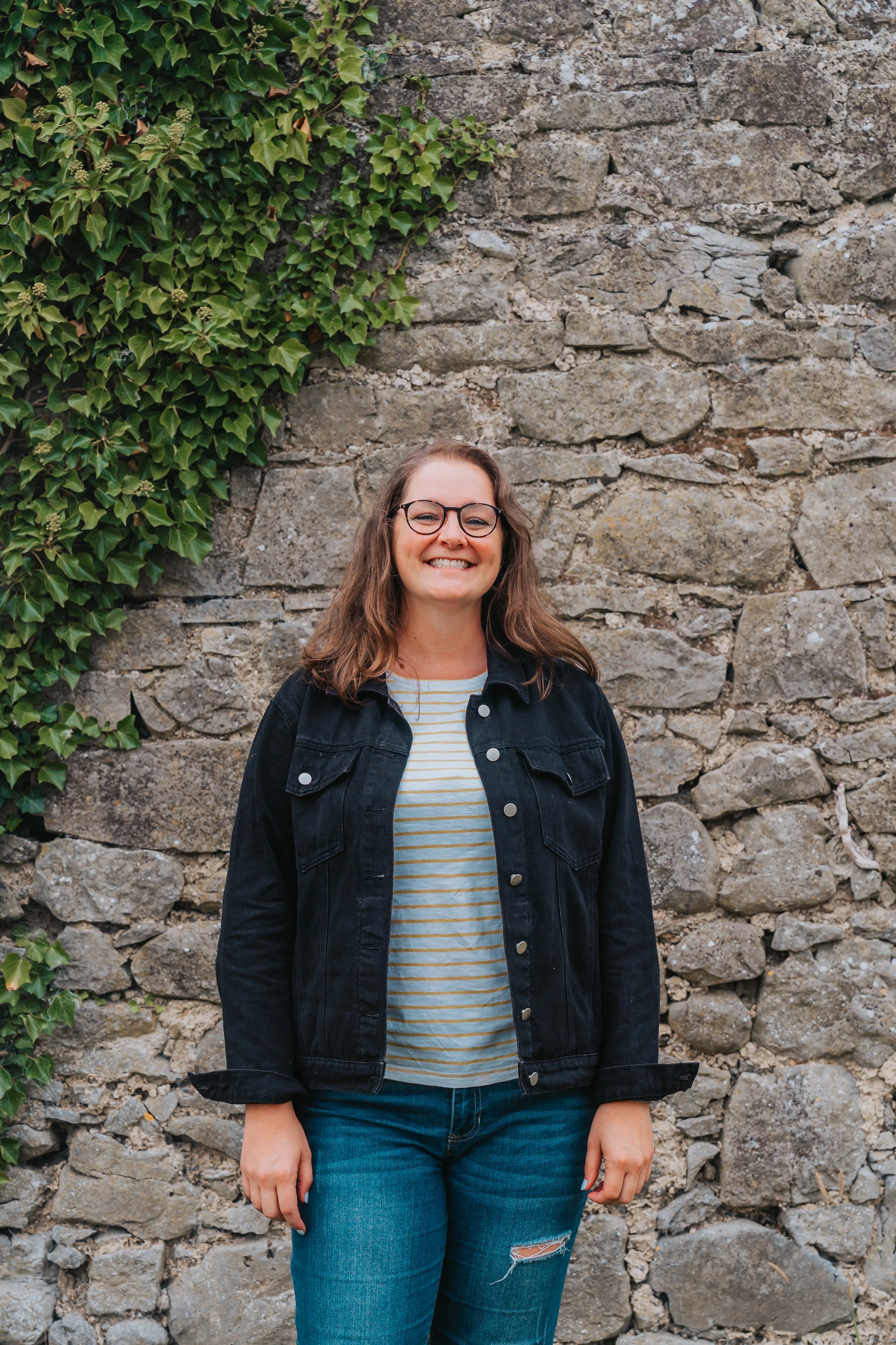 A woman with glasses, curly brown hair, wearing a black jacket over a striped shirt and ripped jeans, standing in front of a stone wall with ivy on the left side.