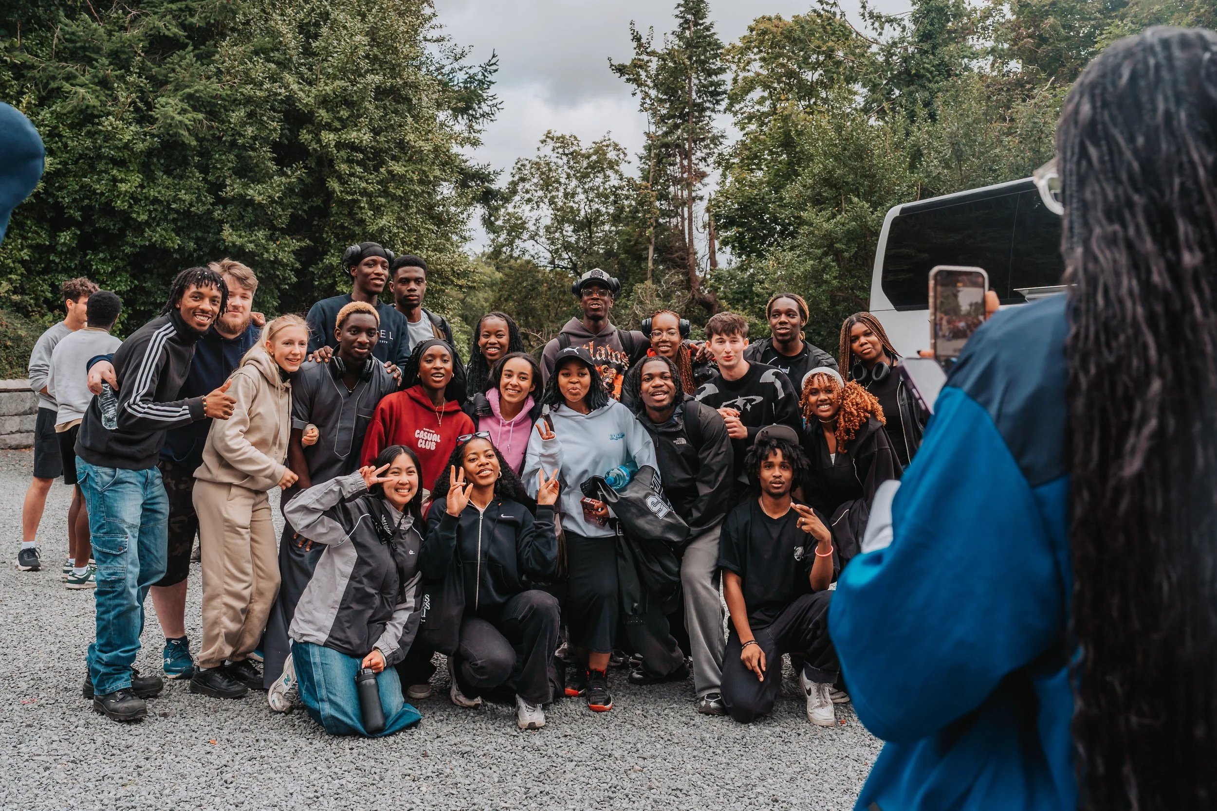 Group of young diverse people gathered outdoors in a forested area, posing for a photo with one person taking the picture.