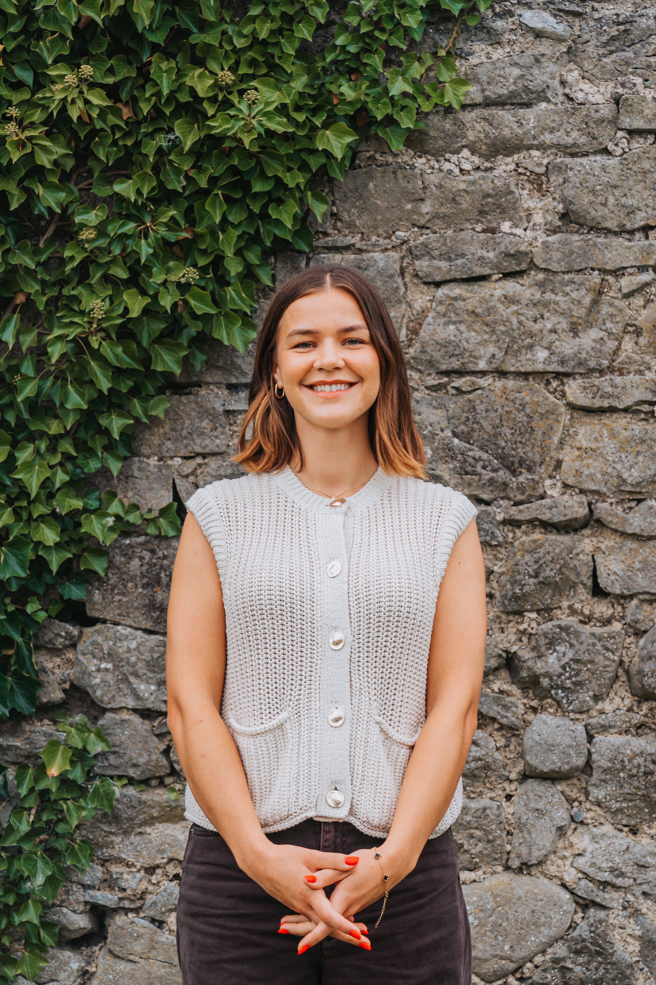 A woman with shoulder-length brown hair smiling at the camera, standing in front of a stone wall with green ivy on the left side.