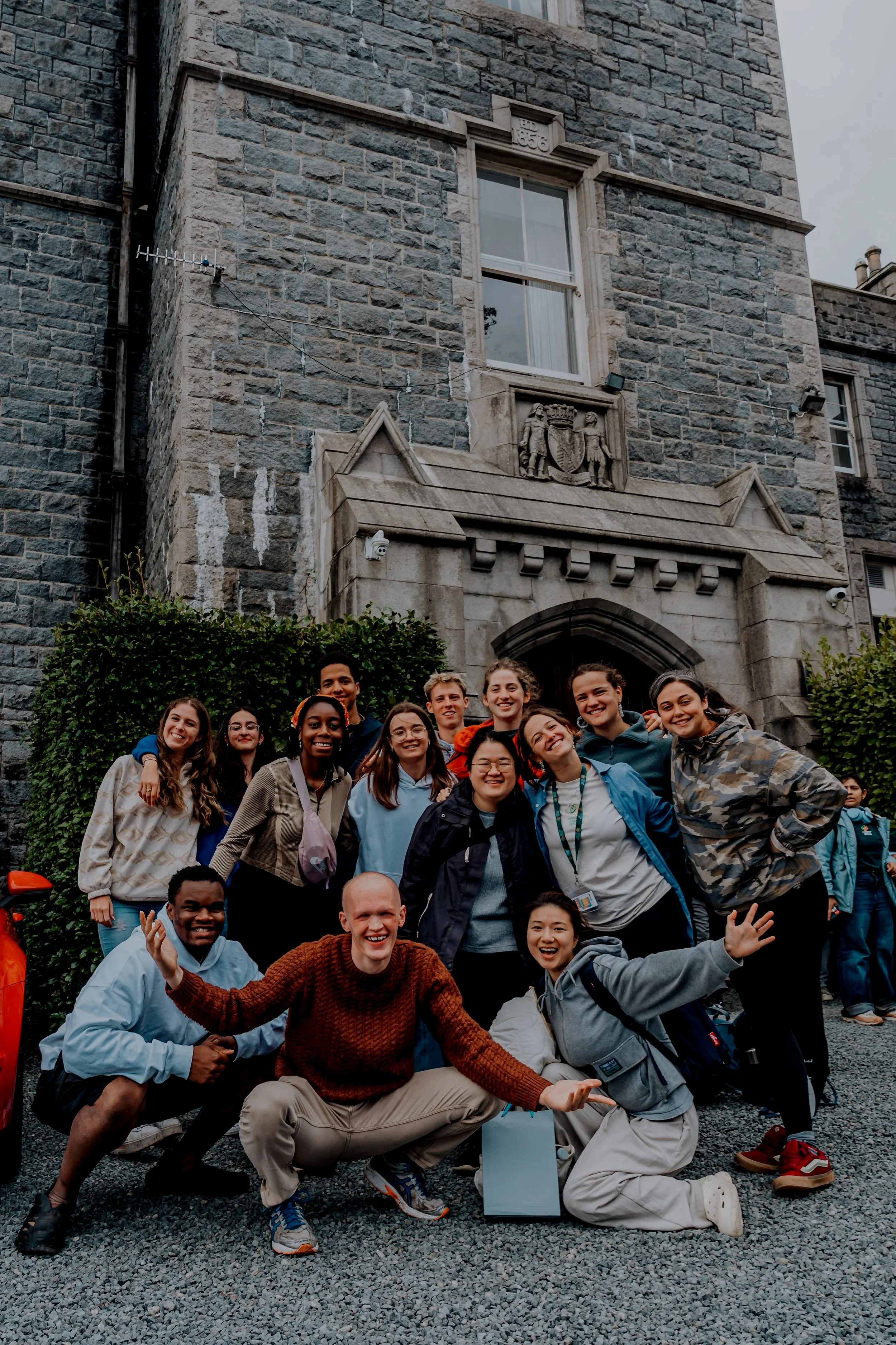 A group of diverse young people smiling and posing for a photo in front of a historic stone building with decorative carvings and windows. Some are standing, while others are kneeling or crouching in the front, showing a lively and cheerful atmosphere.