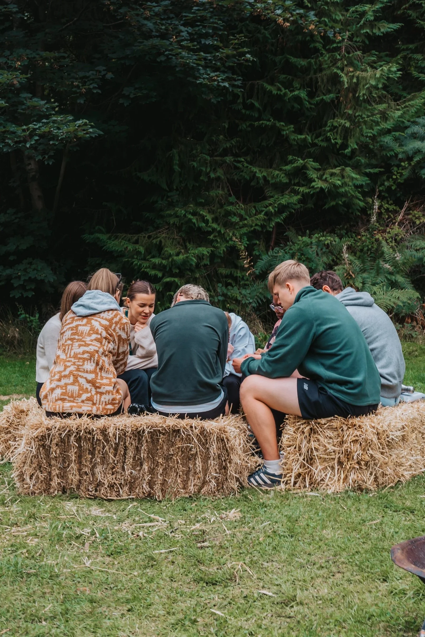 Group of people sitting together in a circle on hay bales outdoors, engaged in conversation, with a backdrop of dense green trees.