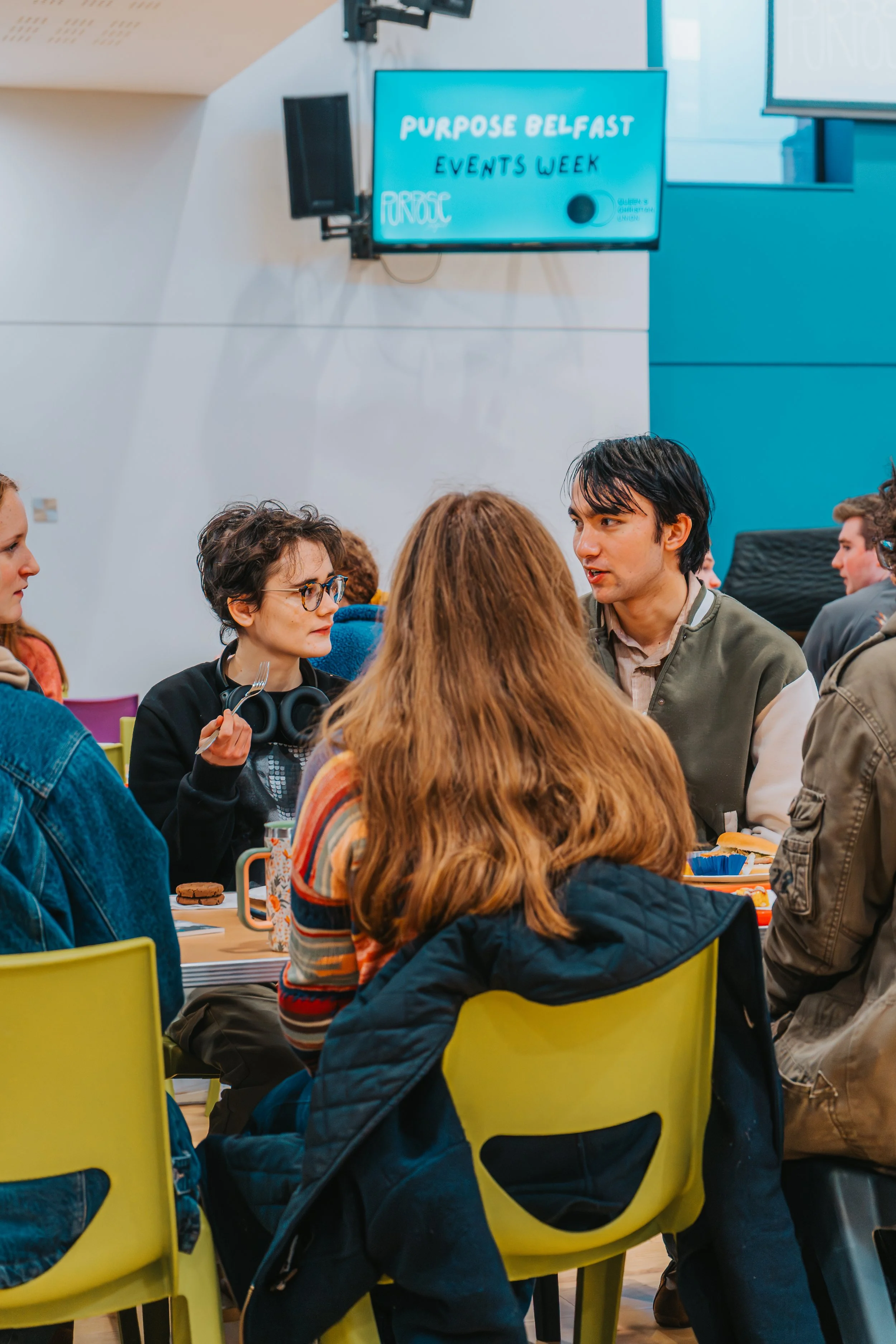 Group of people sitting around a table at an event, with a digital sign in the background reading "Purpose Belfast Events Week."