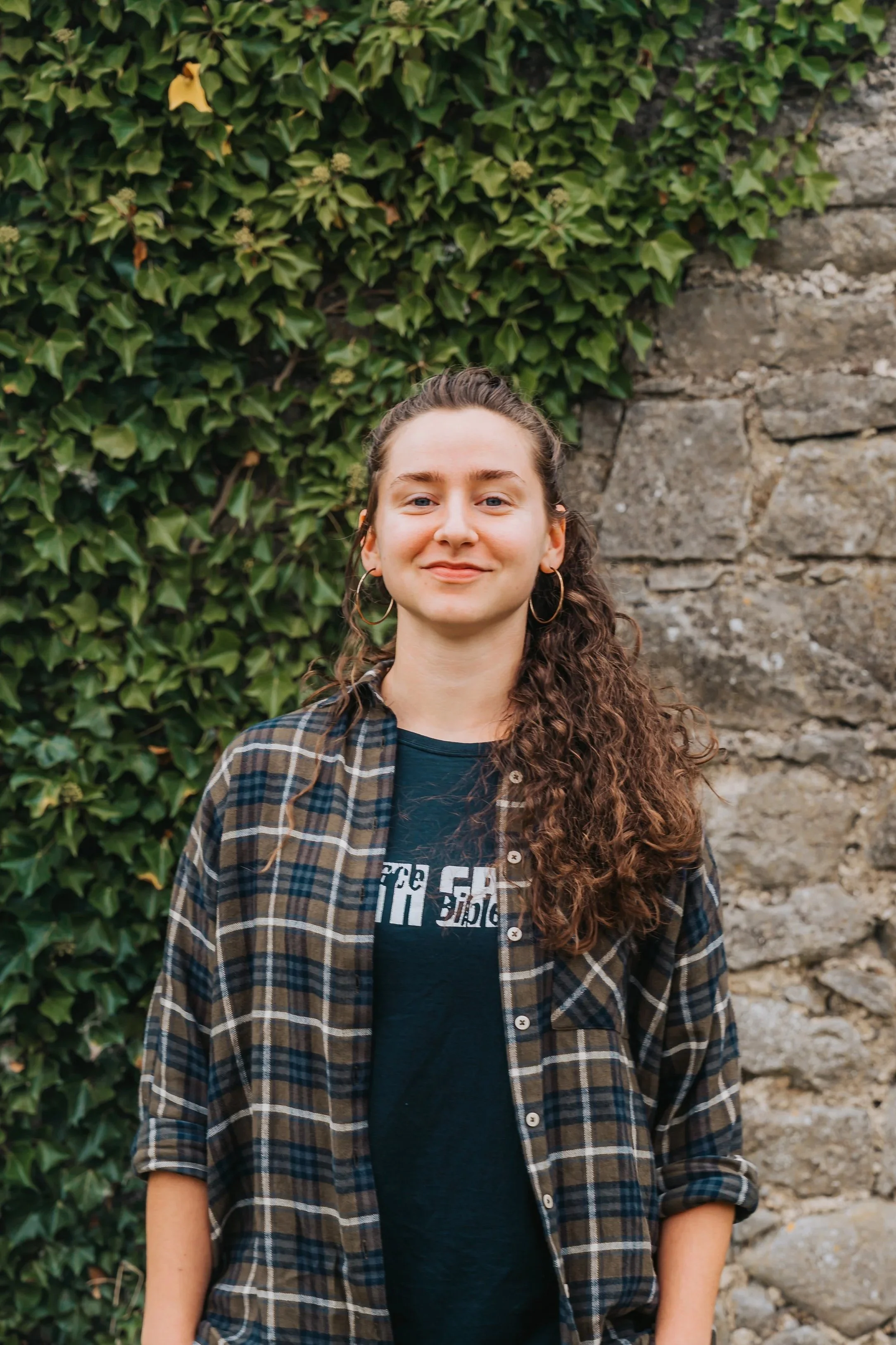 A young woman with curly hair, wearing hoop earrings, a black t-shirt, and an open plaid shirt, standing outdoors in front of a stone wall and green ivy.