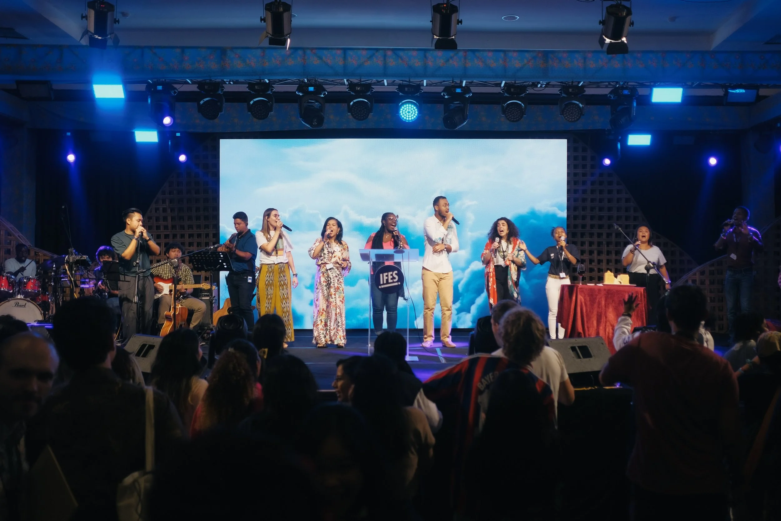 A diverse group of people singing on stage during a concert, with audience members watching in a darkened room.