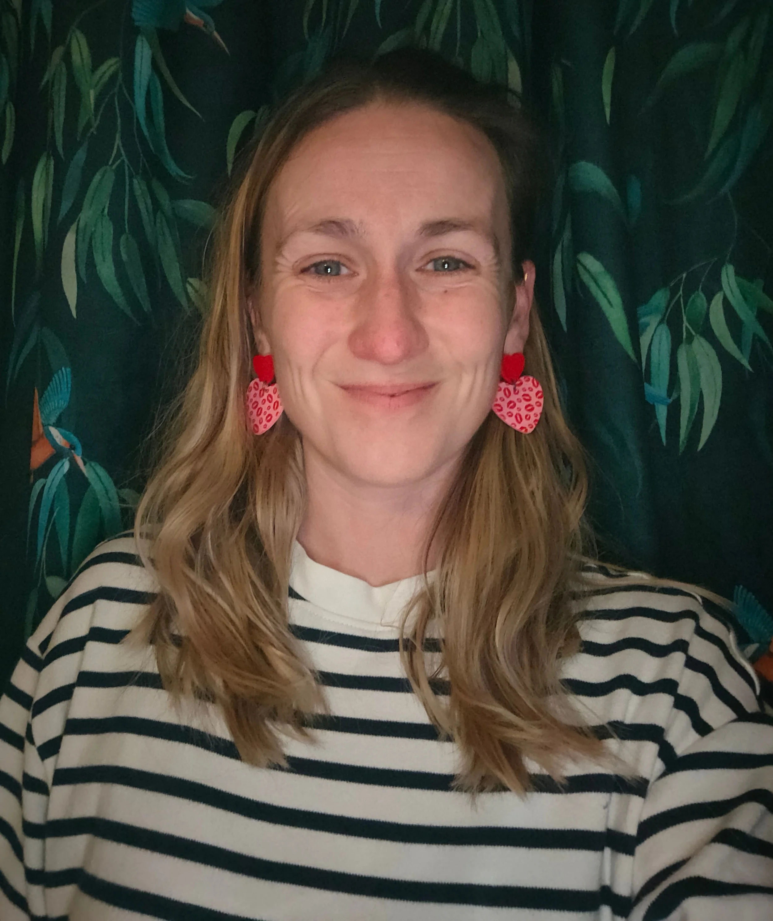 A woman with long, wavy blonde hair wearing a white and navy blue striped top and strawberry-shaped earrings, smiling in front of a leafy green background.