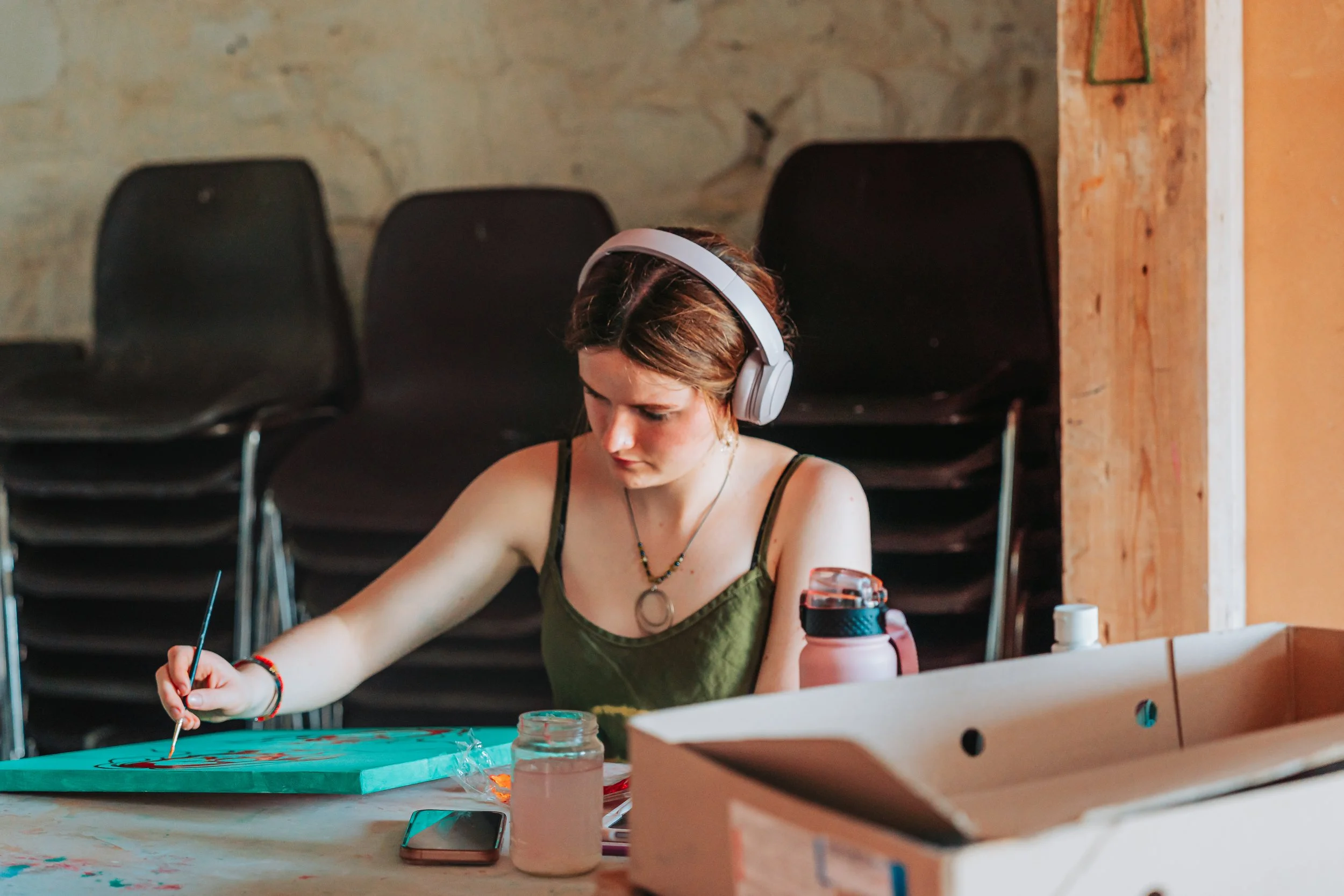 A young woman with short brown hair wearing headphones is painting on a canvas at a table in a rustic space with black chairs and an exposed wall.